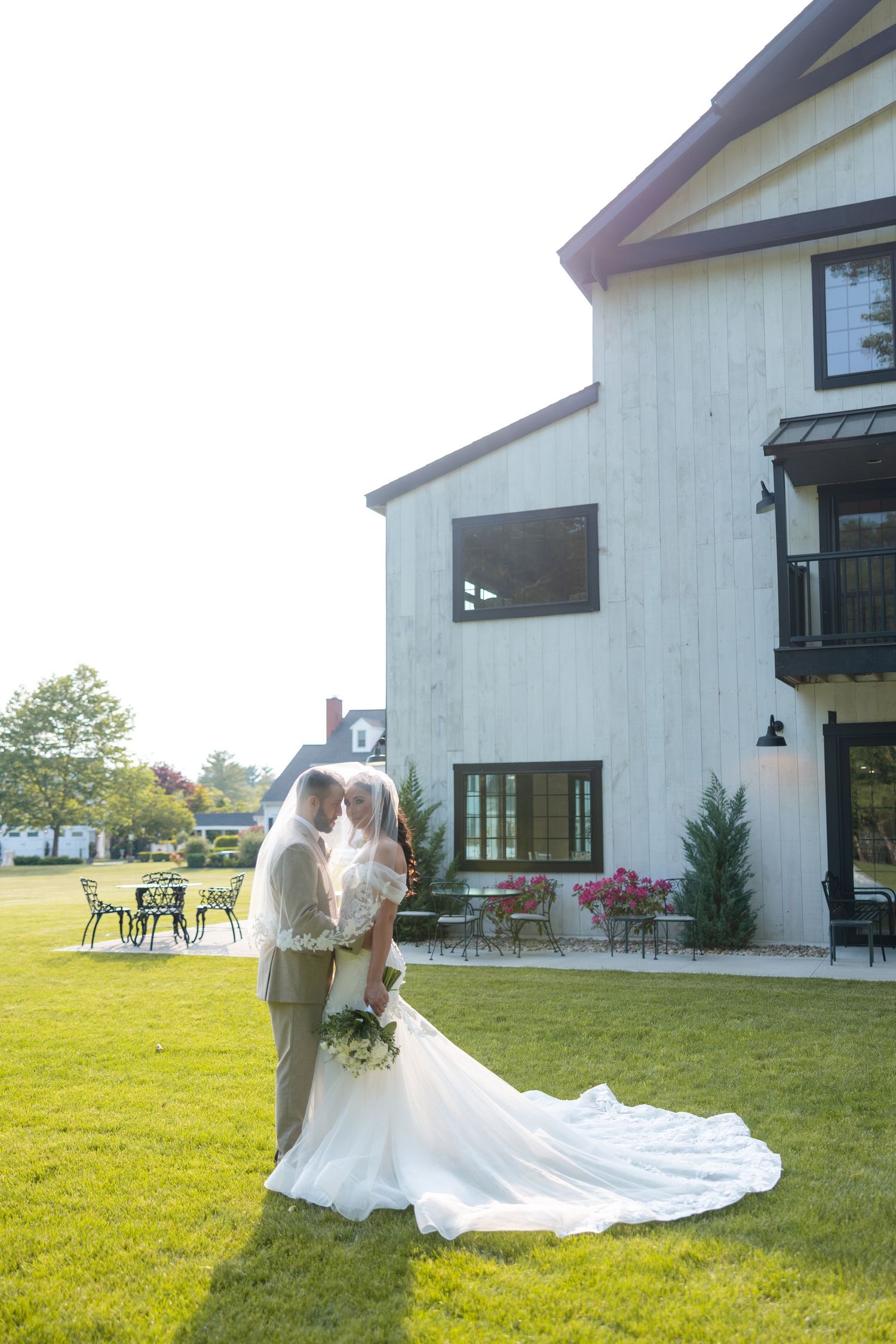A couple in wedding attire stands on a lush green lawn in front of a modern white farmhouse on a sunny day.