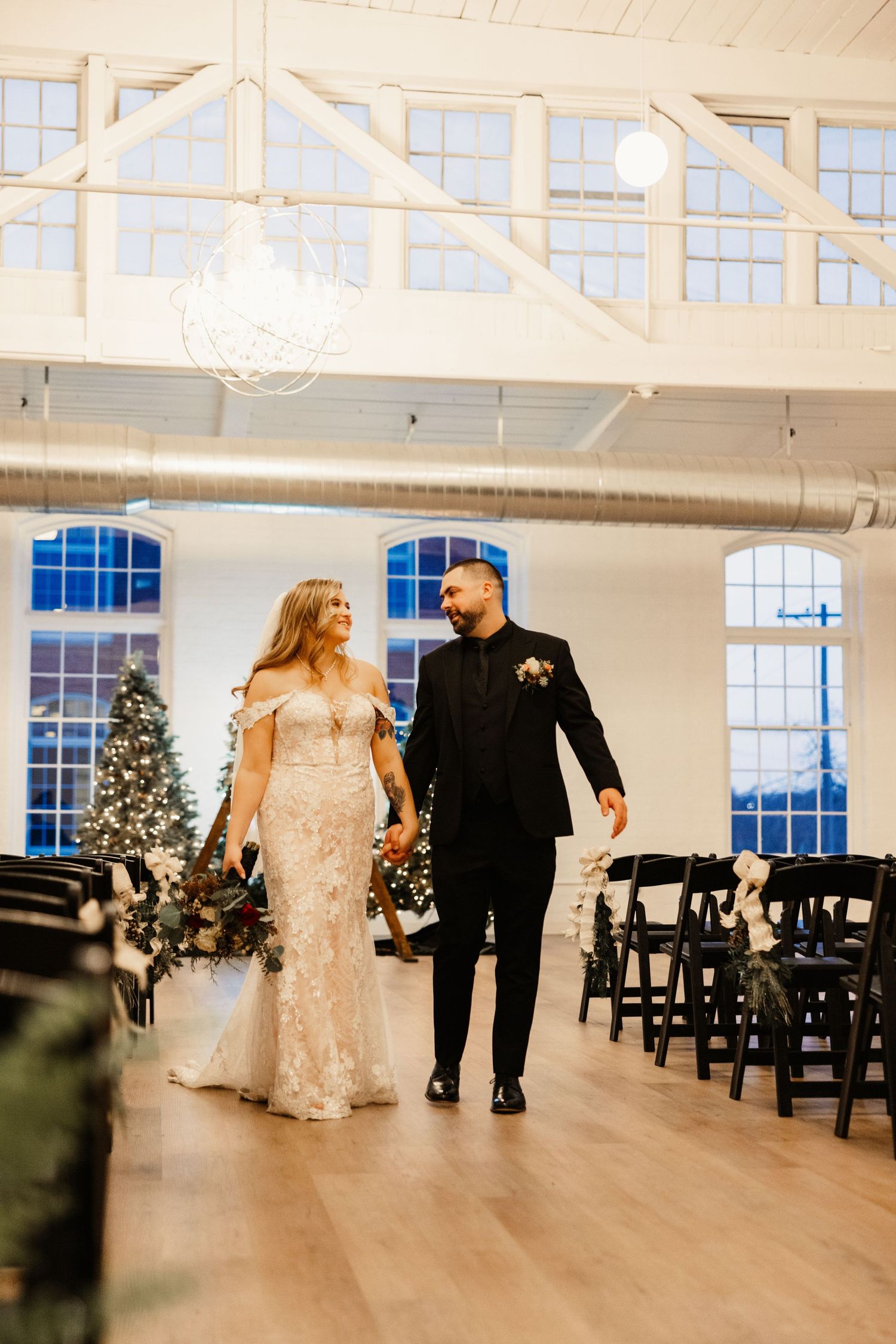 A bride and groom walk hand in hand through an elegant venue with Christmas trees and chandeliers.