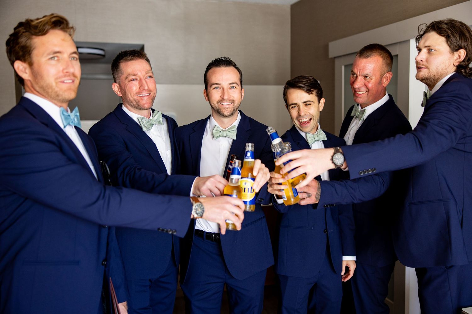 Groomsmen in navy suits toast with beer bottles during pre-wedding celebration.