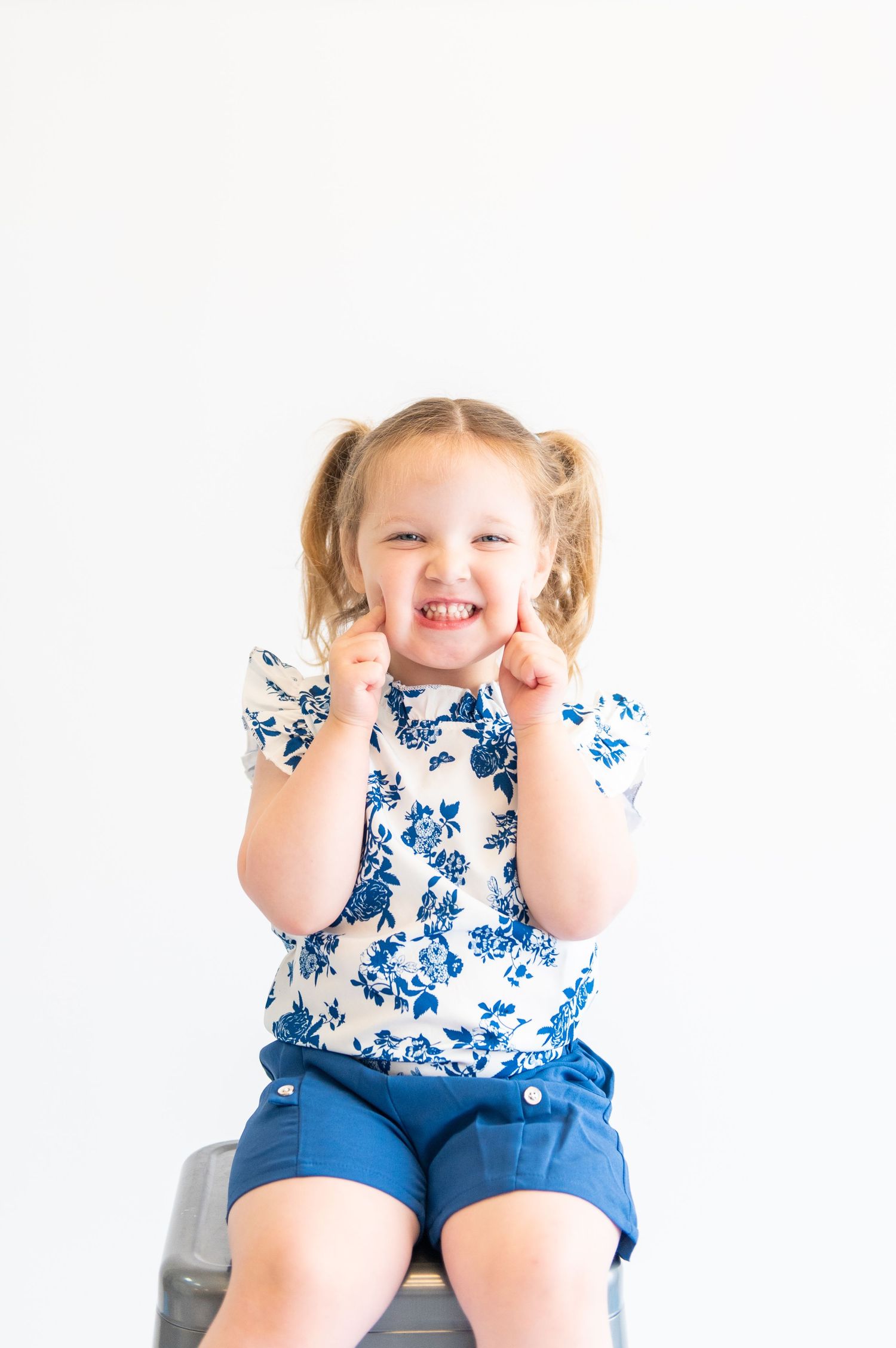 Child in floral print top and blue shorts sitting on stool with excited expression.