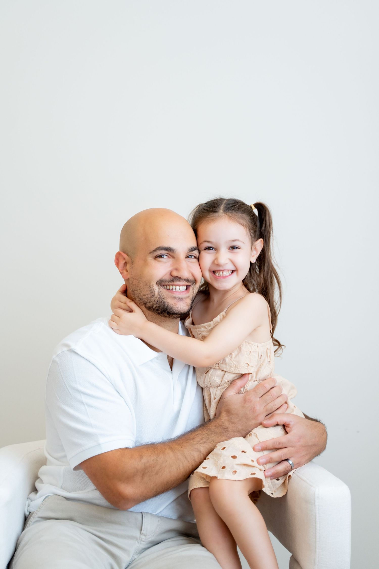 Father and daughter hugging and smiling while sitting in white chair against plain wall.