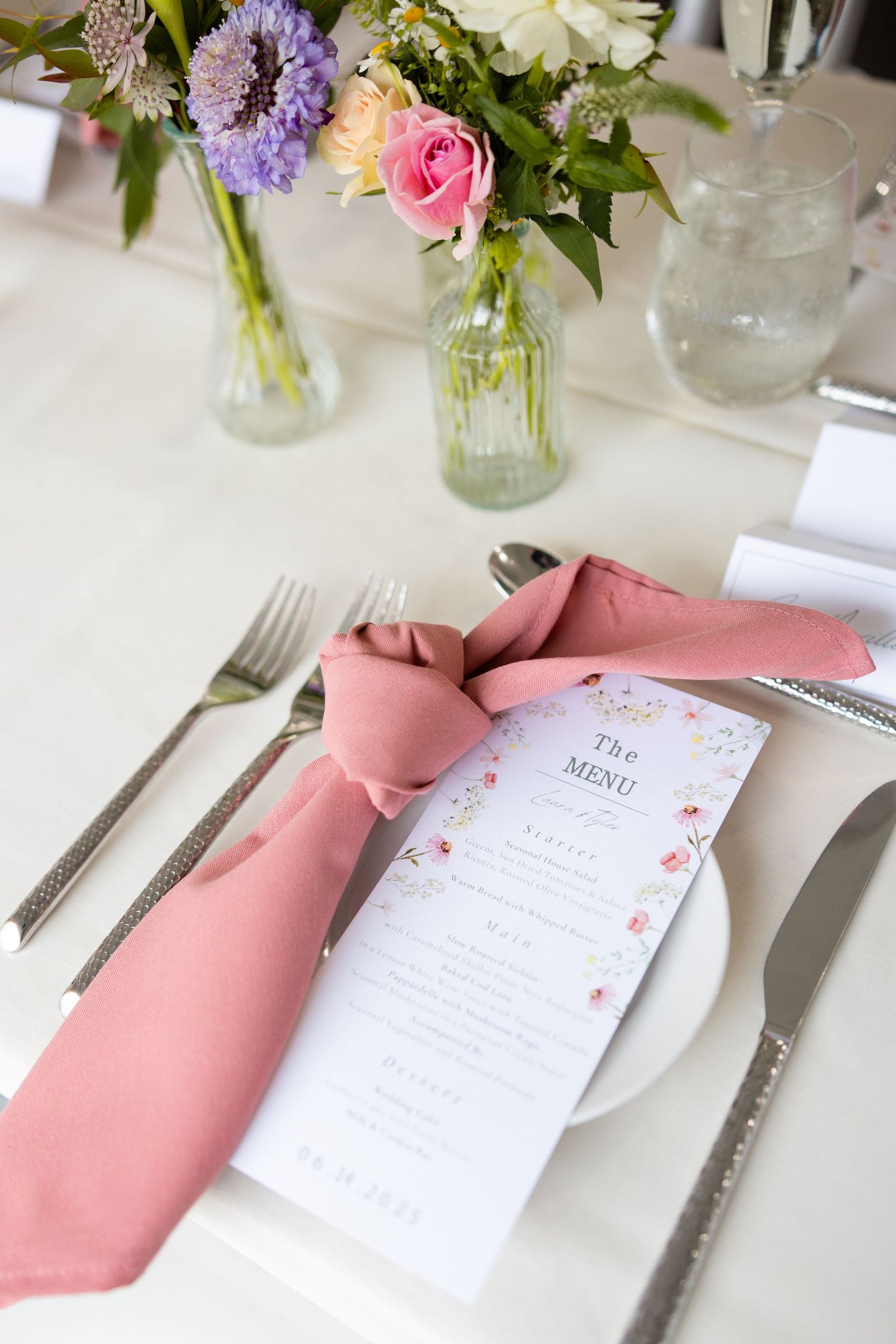 Wedding reception place setting with pink napkin, floral menu card and centerpiece of fresh flowers in glass vases.