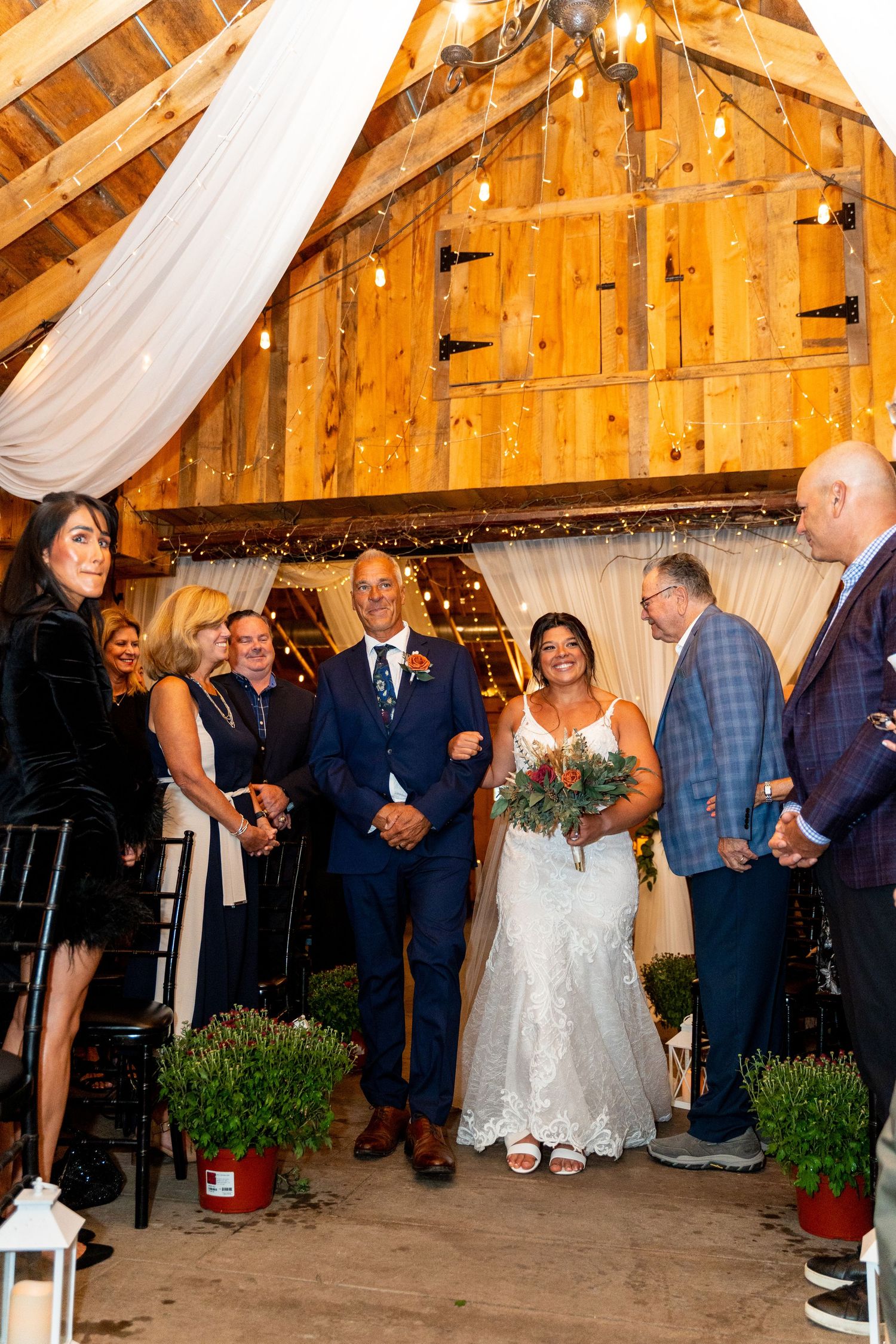 Wedding ceremony in rustic wooden barn decorated with white fabric and string lights.