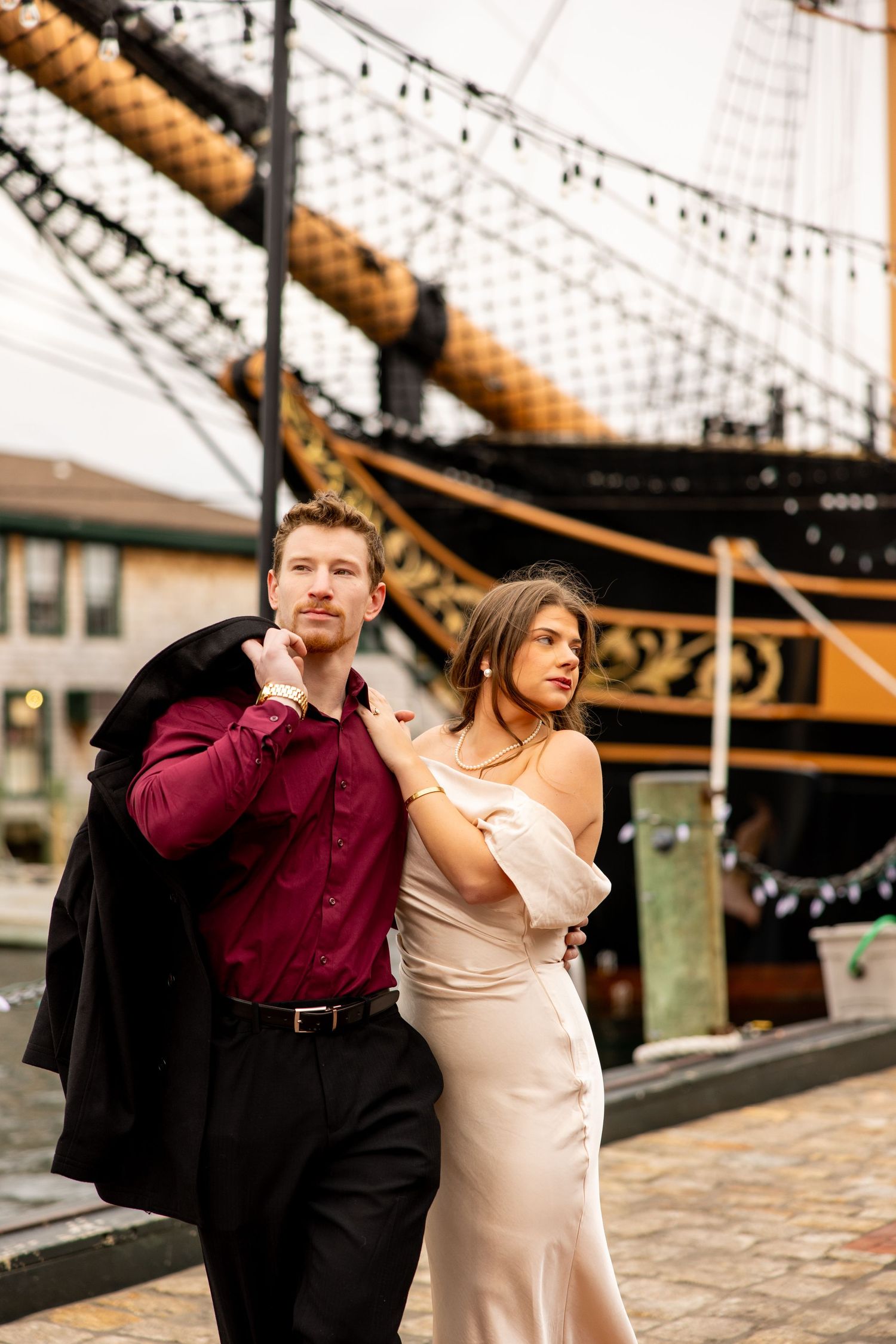 A couple in formal attire poses in front of a historic ship with string lights and rigging visible in the background.