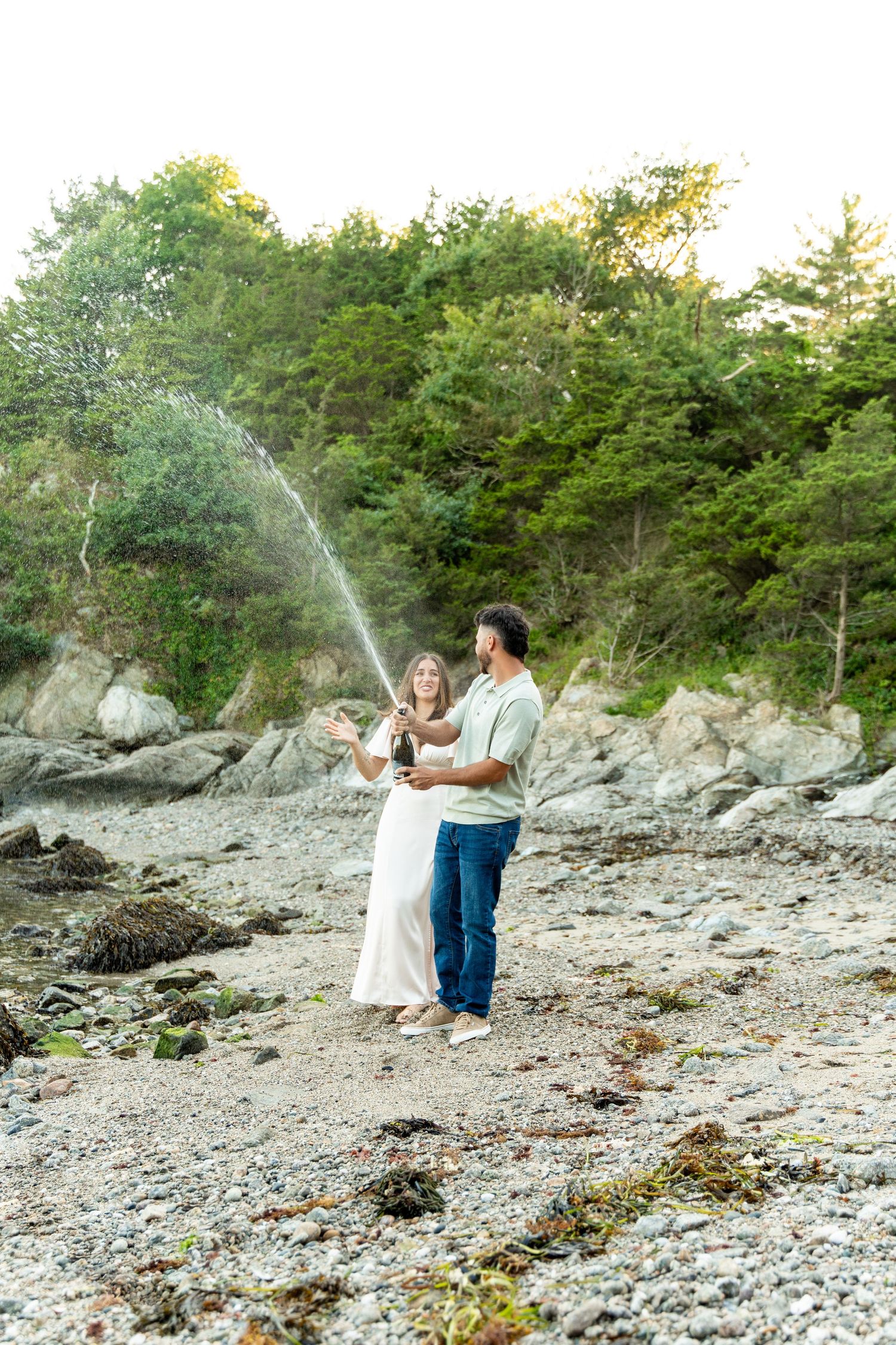 Playful couple celebrating with champagne spray on rocky beach surrounded by trees.