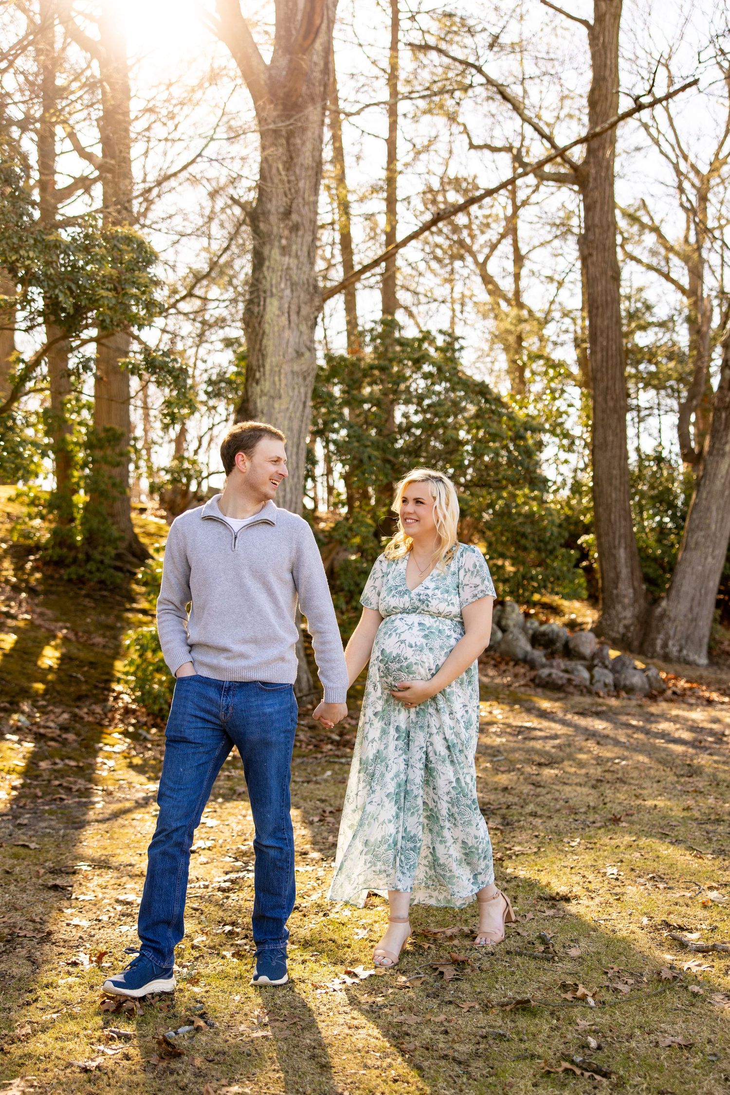 A couple holds hands while walking through a sun-dappled wooded area.