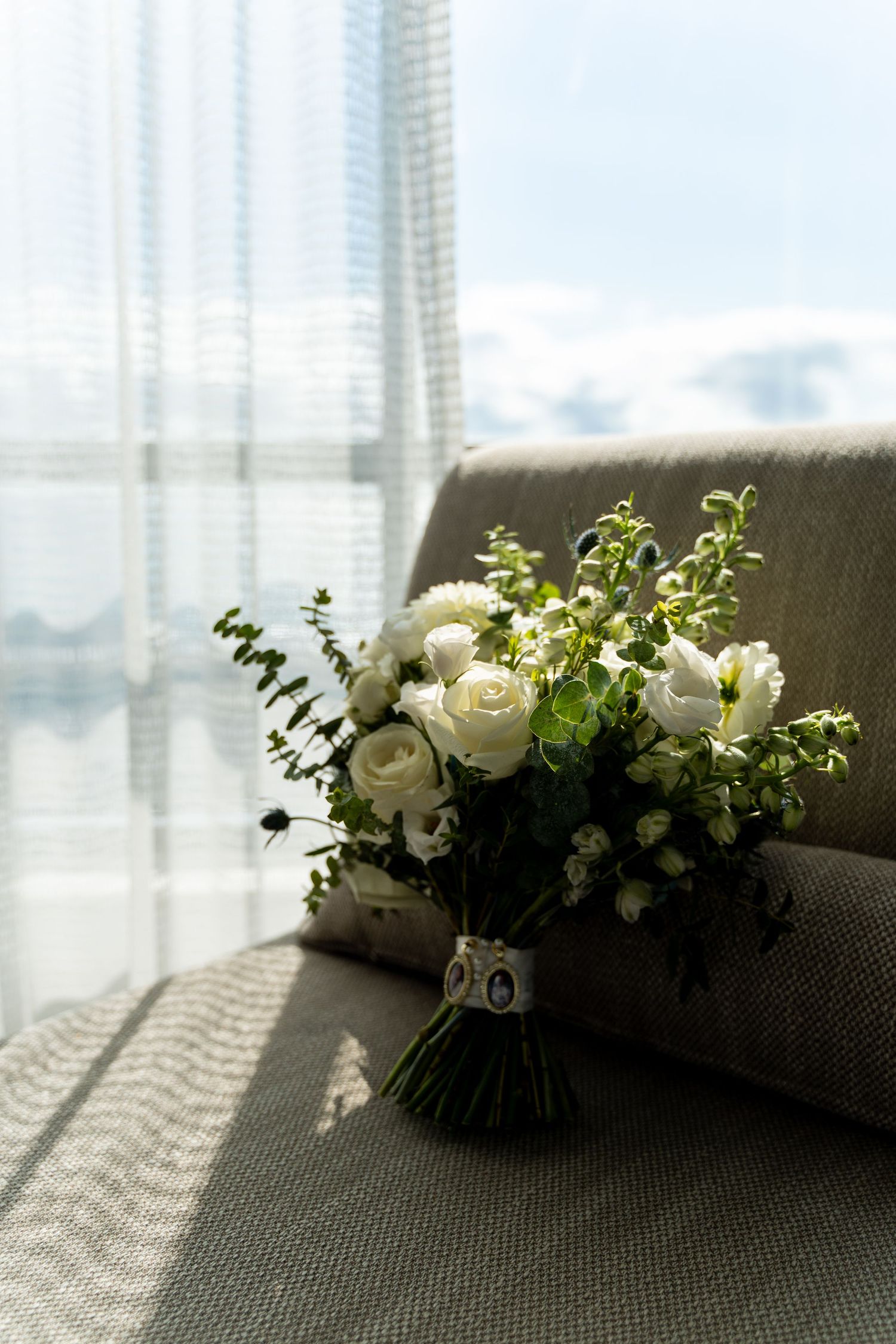 White rose bouquet with greenery resting on gray couch by window.