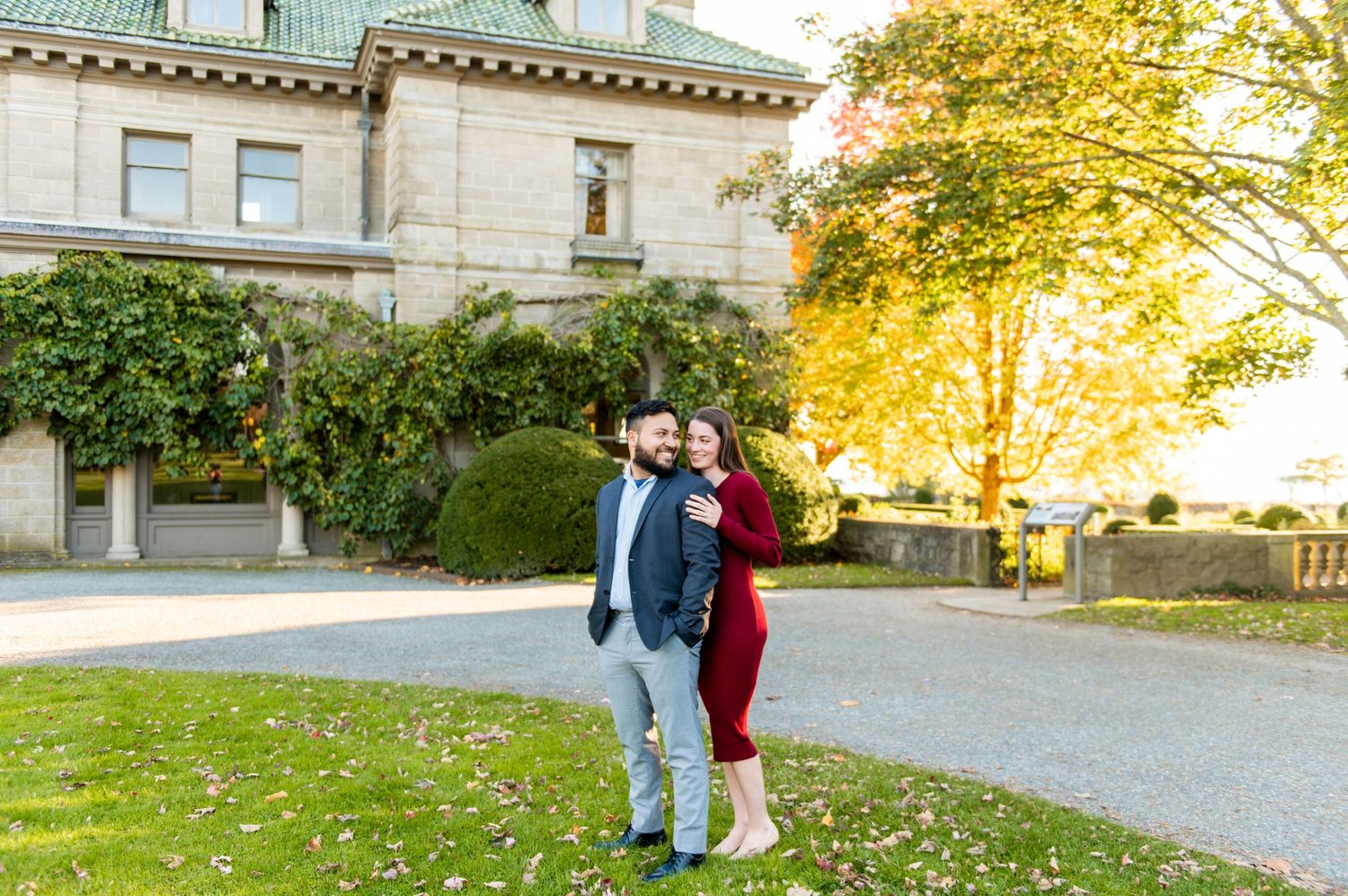 A couple stands together on a gravel path in front of a grand stone mansion.