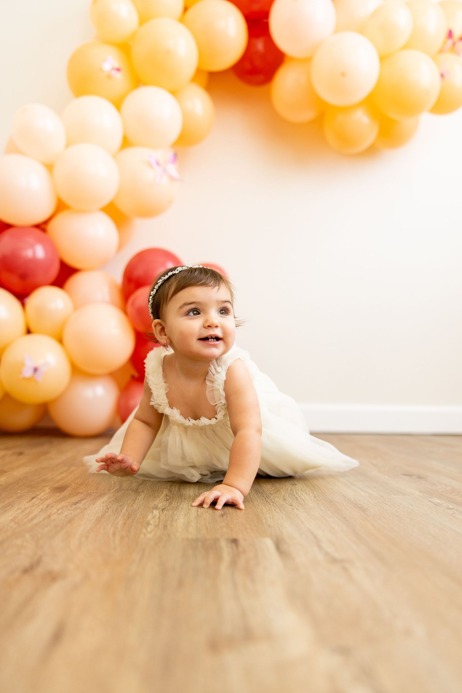 Baby in tulle dress crawls on wooden floor with balloon decorations.