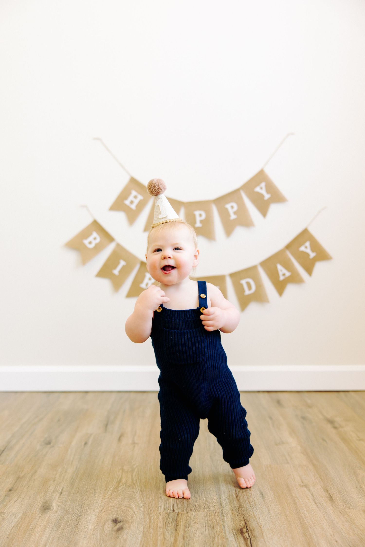 Baby in navy overalls standing under Happy Birthday banner on wooden floor.