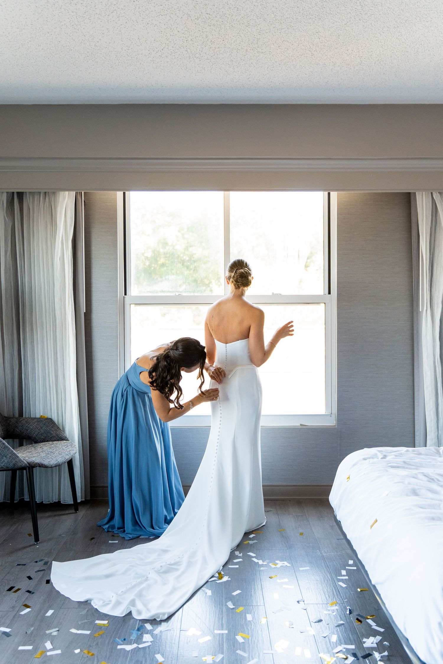 Bridesmaid in blue dress helps bride with wedding gown near bright window.