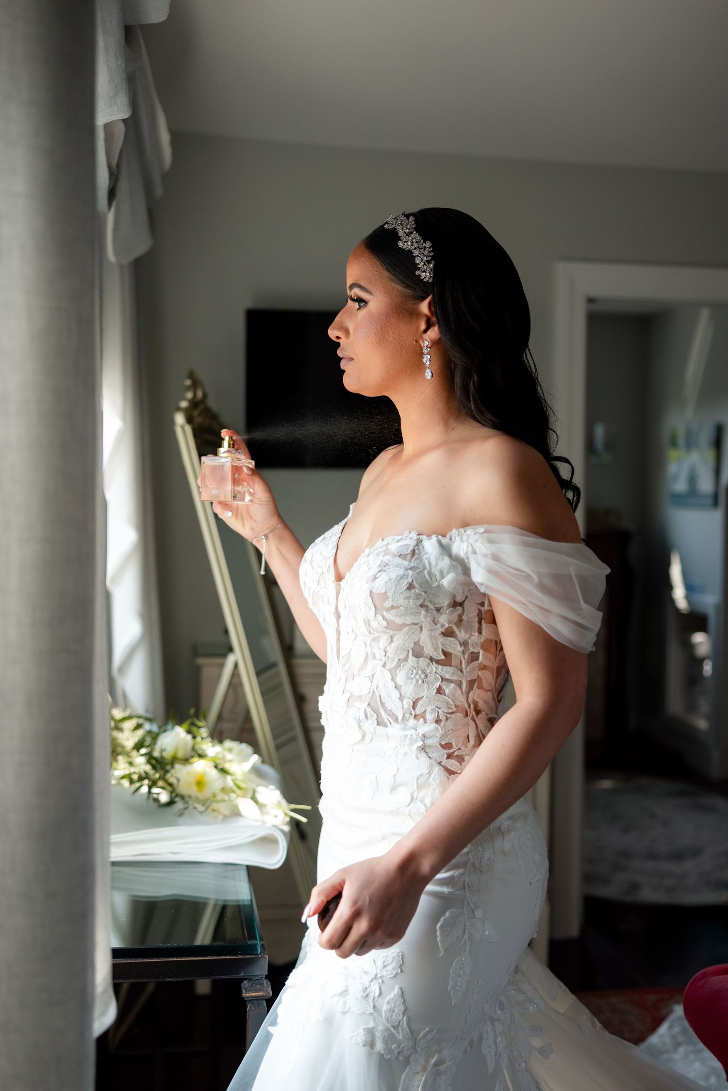 A bride in an off-shoulder white lace wedding gown with a beaded headpiece stands in profile near a window.