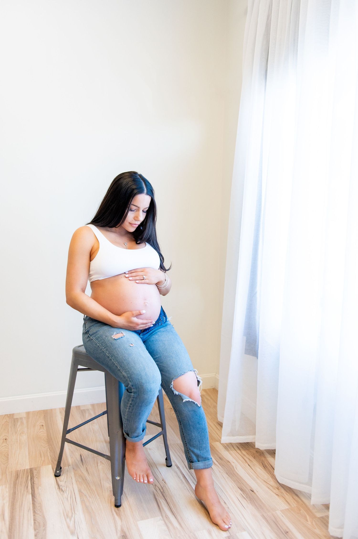 Expectant mother in casual attire sitting on metal stool by window in minimalist setting.