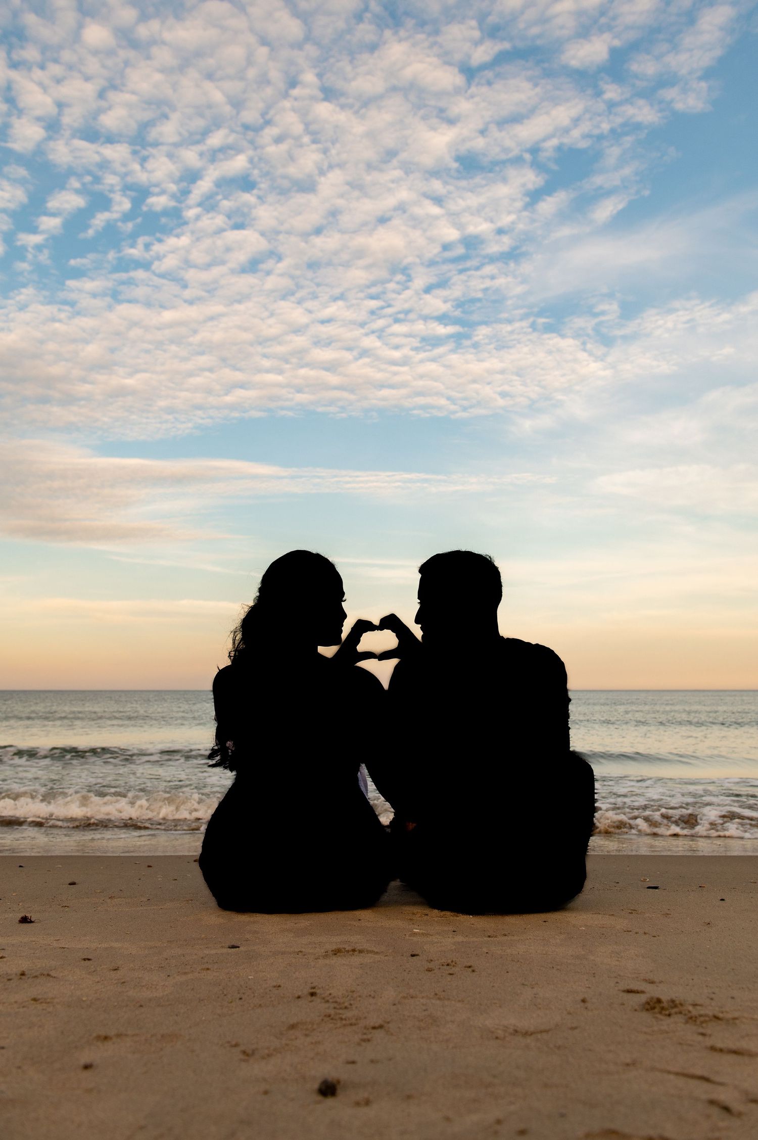 Silhouette of two people making heart shape with hands while sitting on beach at sunset.