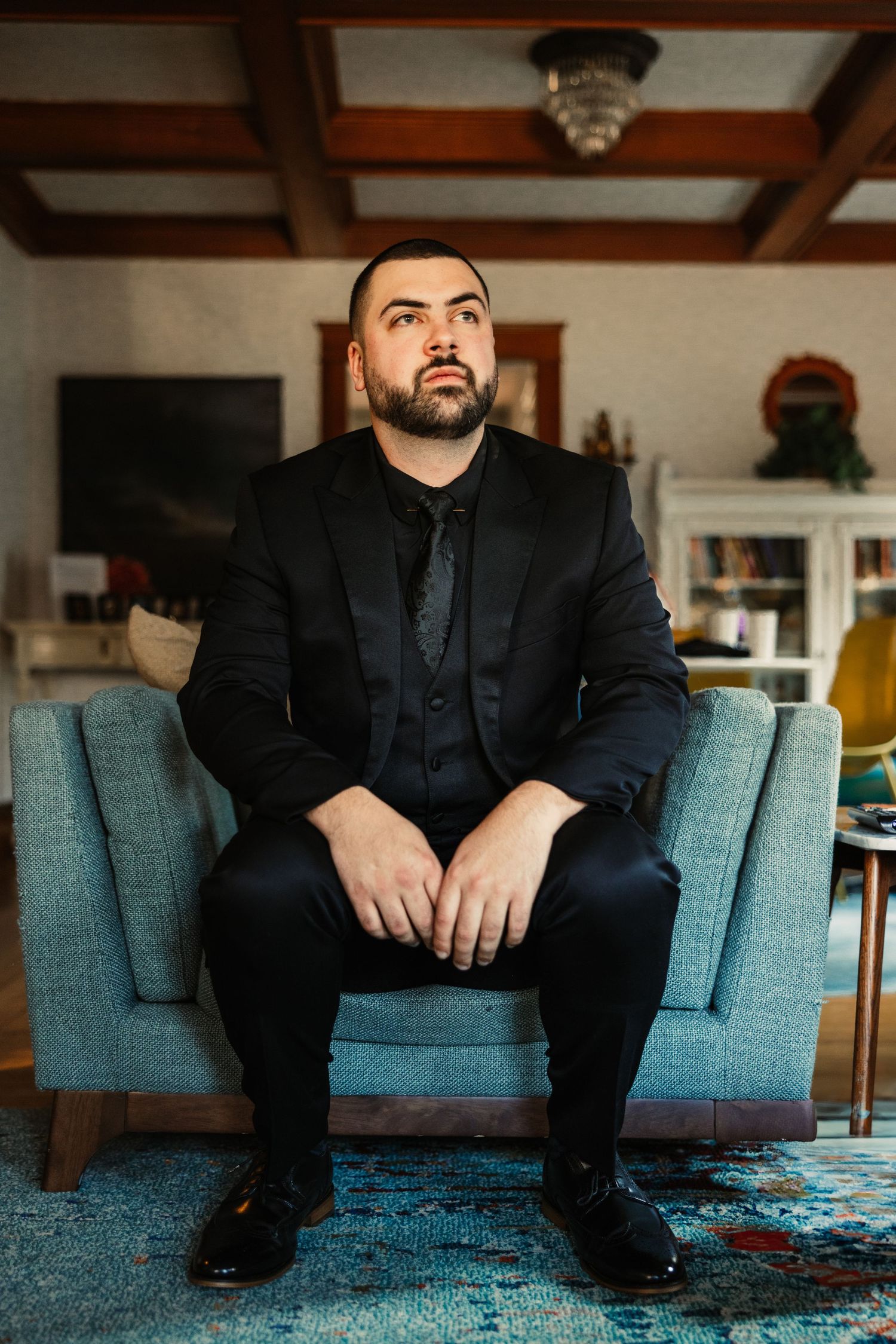 Man in black suit sitting on blue couch in living room setting.