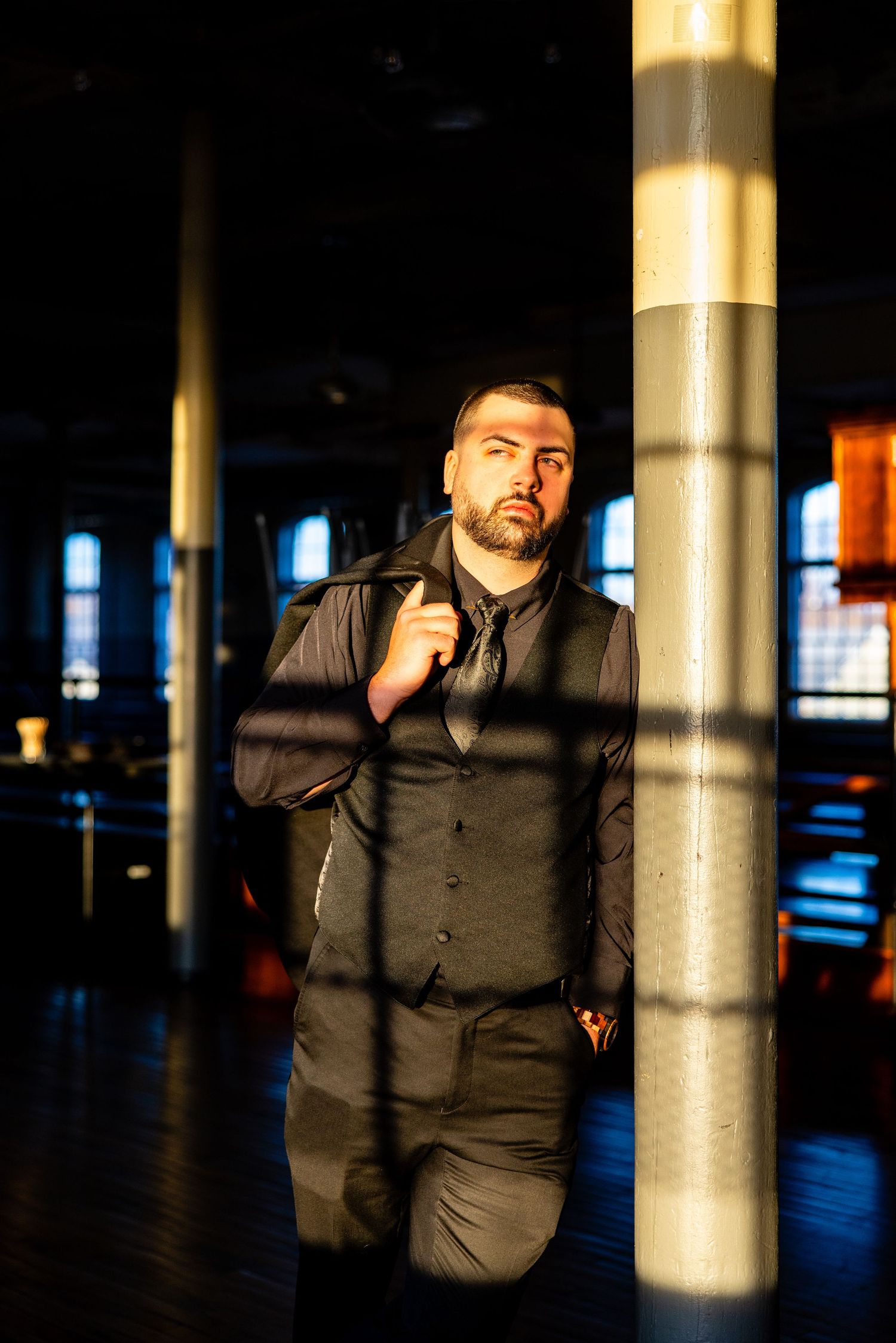 Dramatic portrait of groom in dark suit standing by illuminated pillar in moody venue.