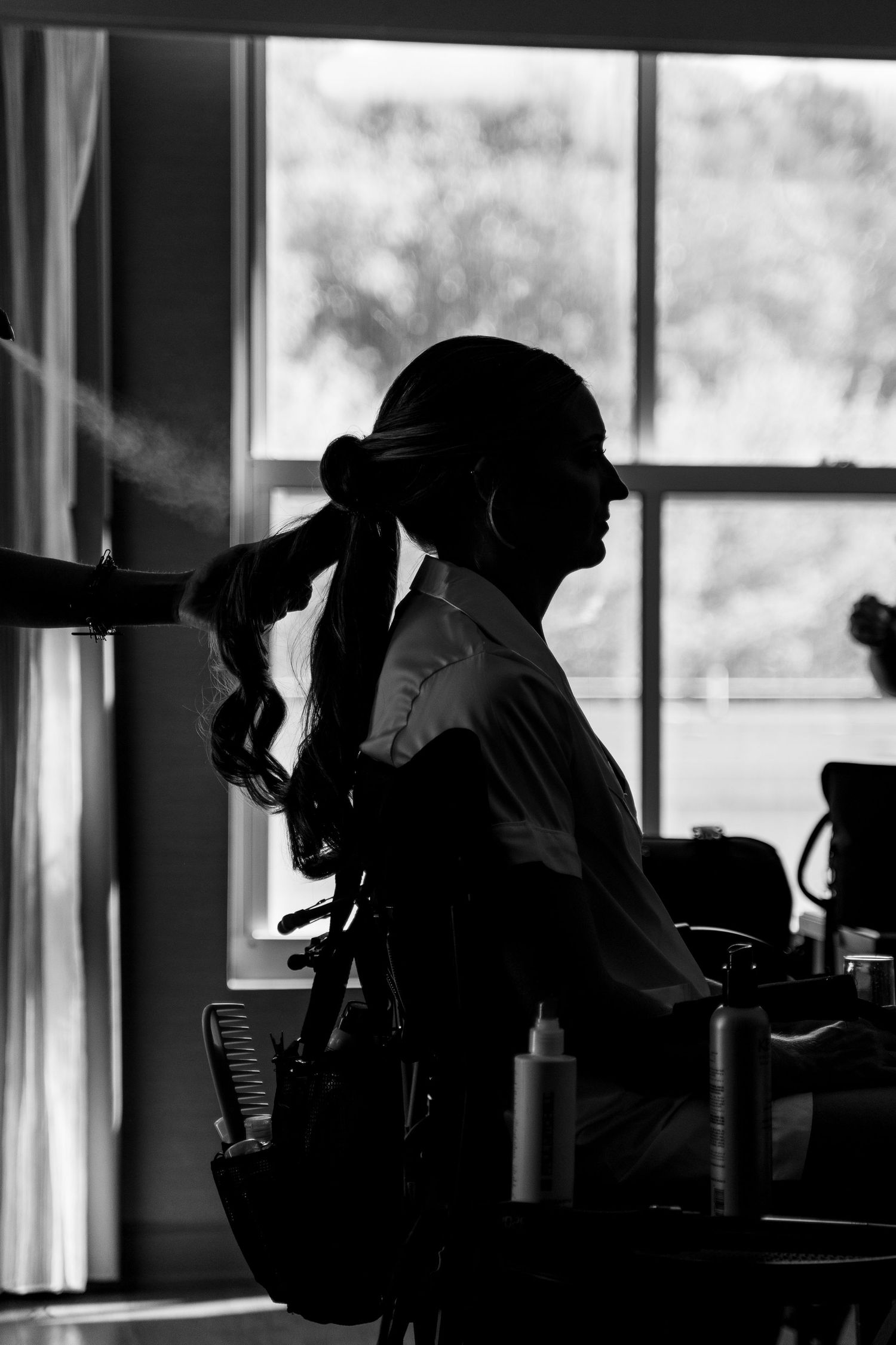 Silhouette of a hairstylist working on bride's hair near a bright window.