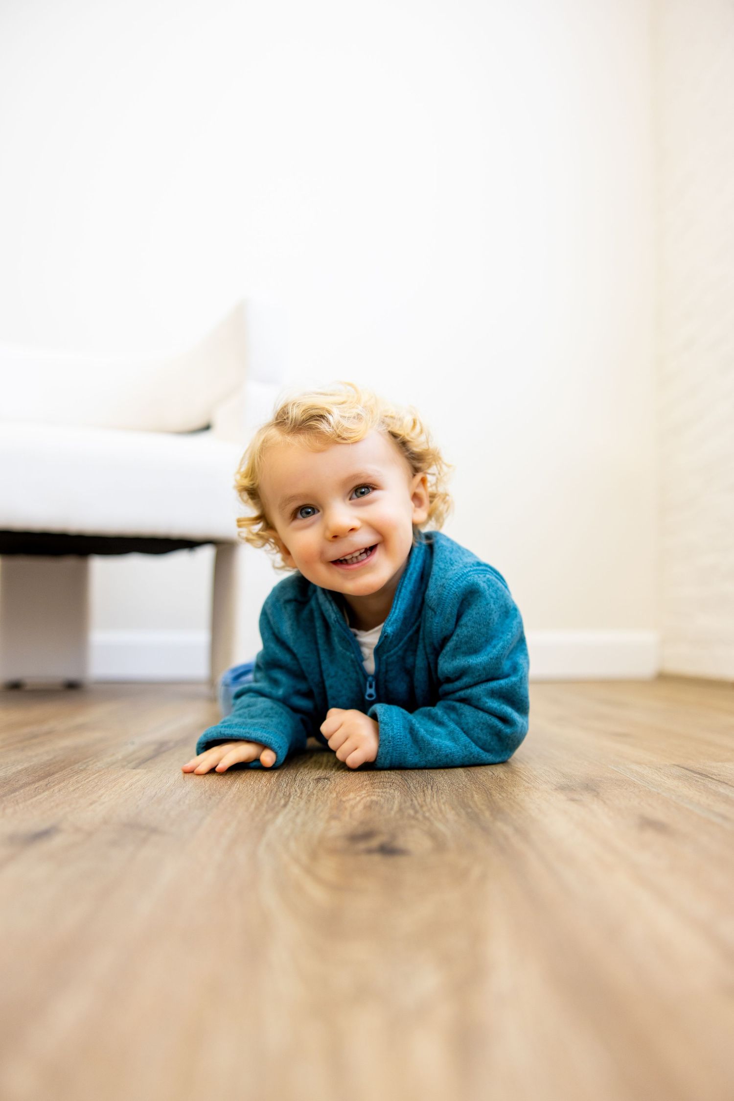 Cheerful toddler in blue sweater lying on hardwood floor.