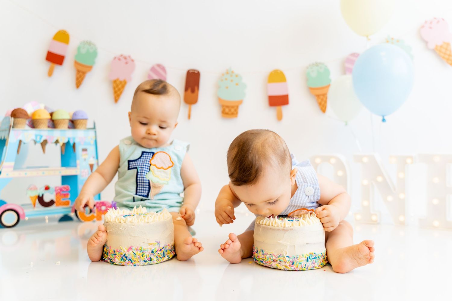 Two babies sitting with matching birthday cakes against an ice cream themed backdrop.