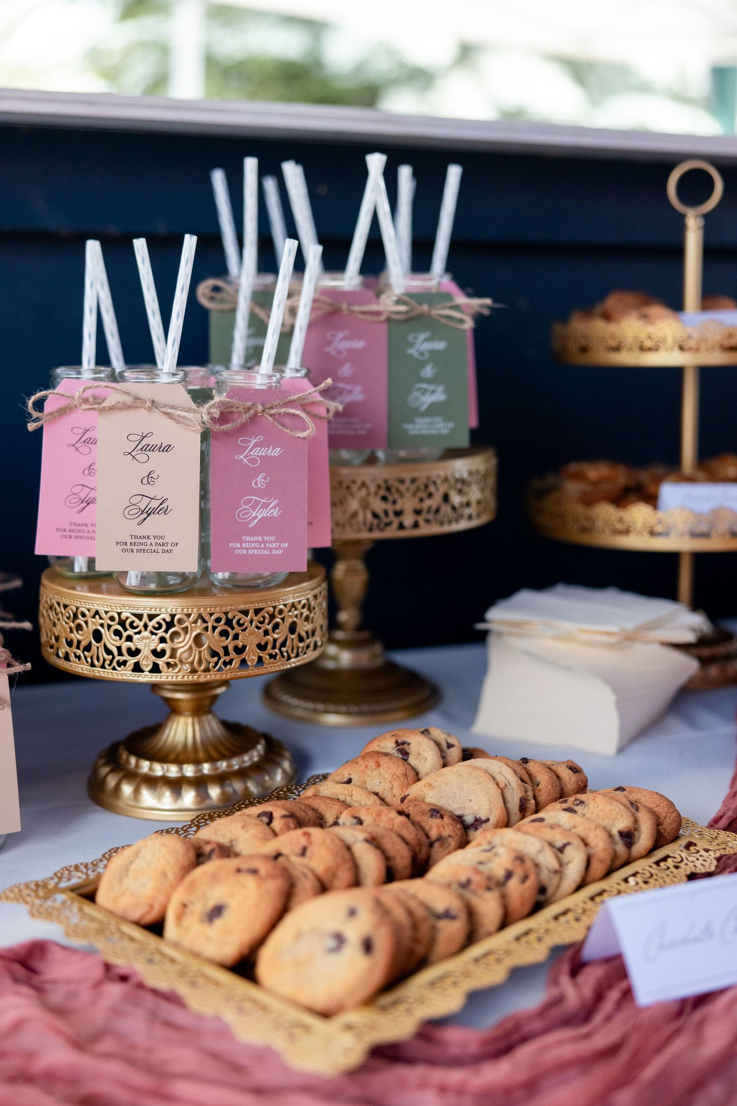 Elegant dessert table display with chocolate chip cookies and pink mason jar drinks on ornate gold pedestals.