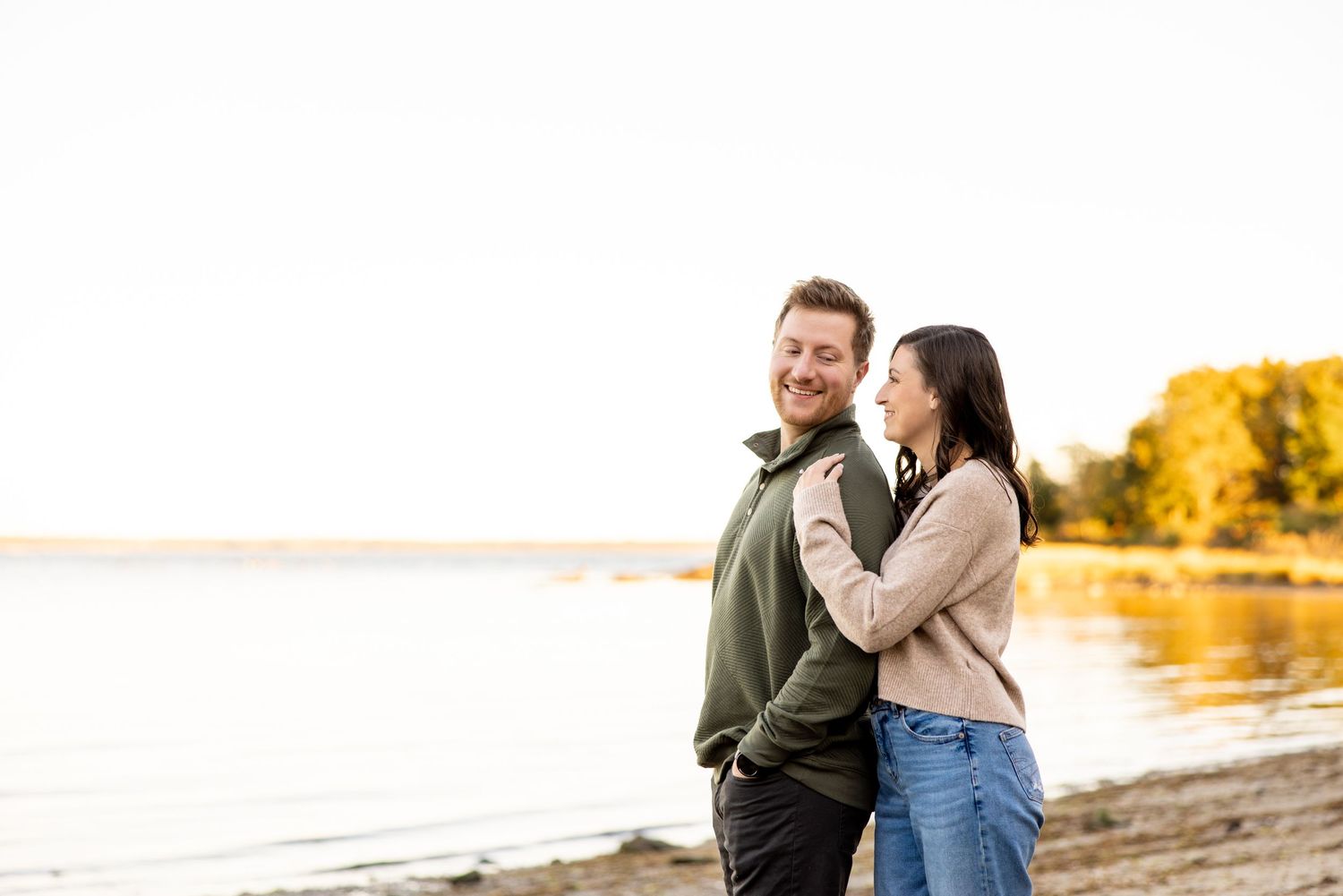 A couple in casual wear shares a tender moment by the water at sunset.