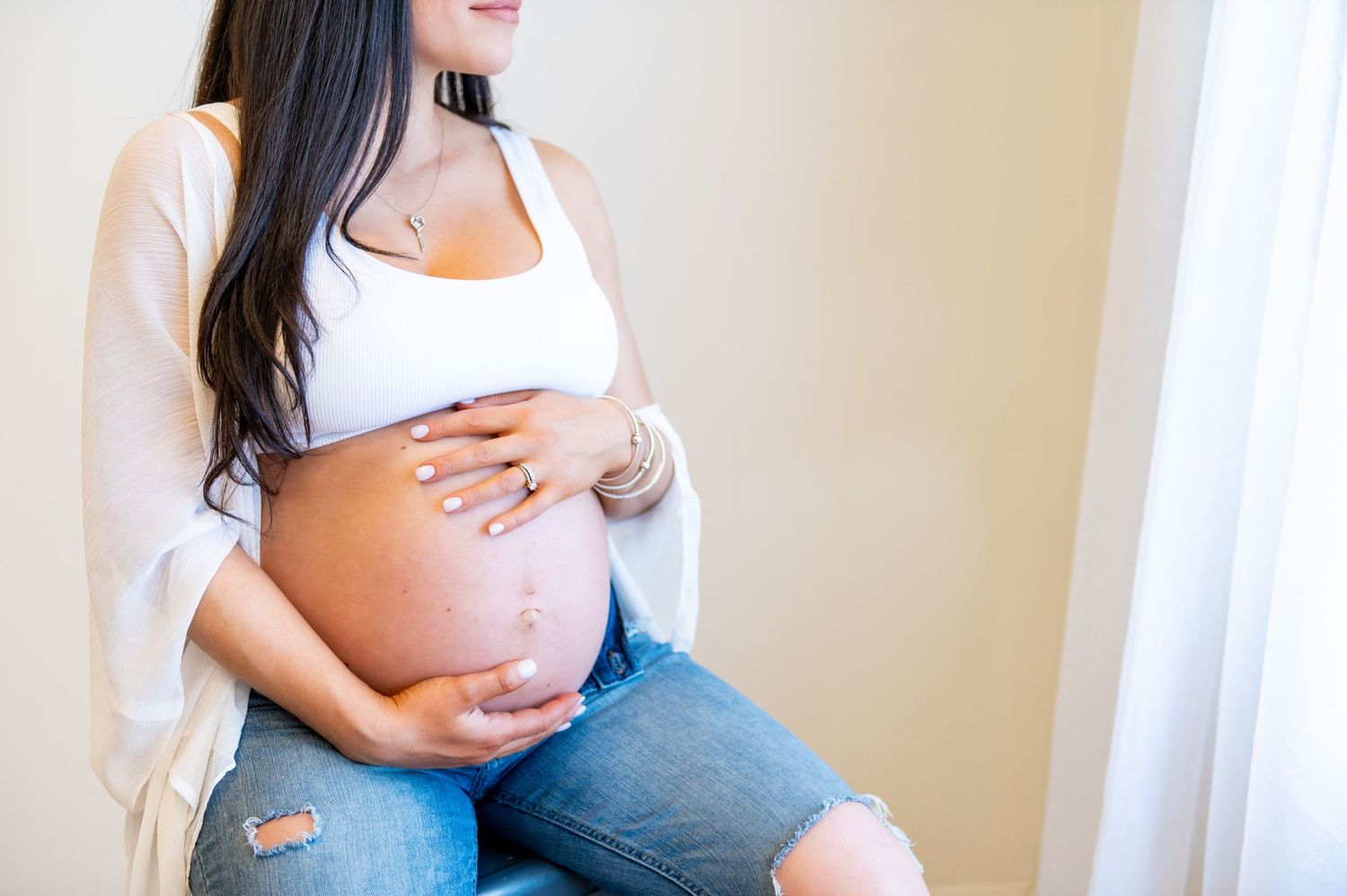 A pregnant person wearing a white tank top and jeans cradles their baby bump while sitting near a window.