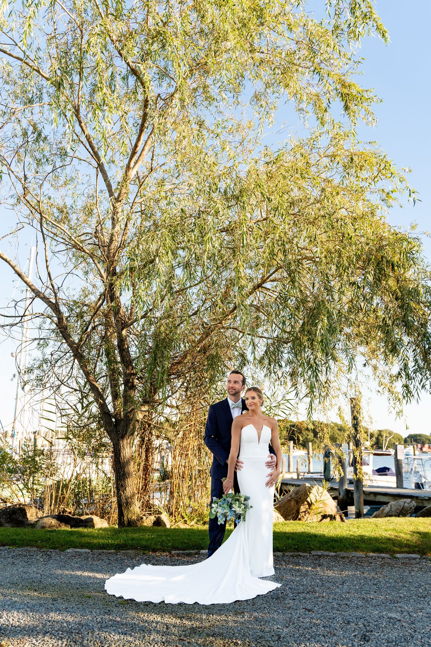 A wedding couple poses beneath a weeping willow tree on a waterfront property.