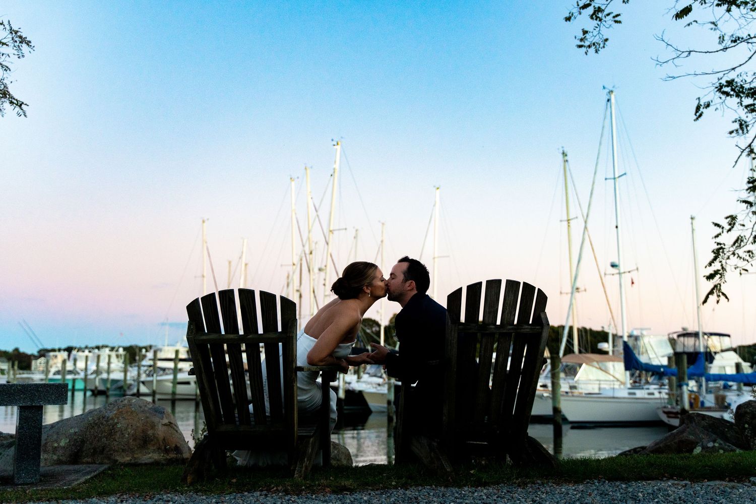 Romantic silhouette of couple sharing moment in Adirondack chairs by marina at sunset.