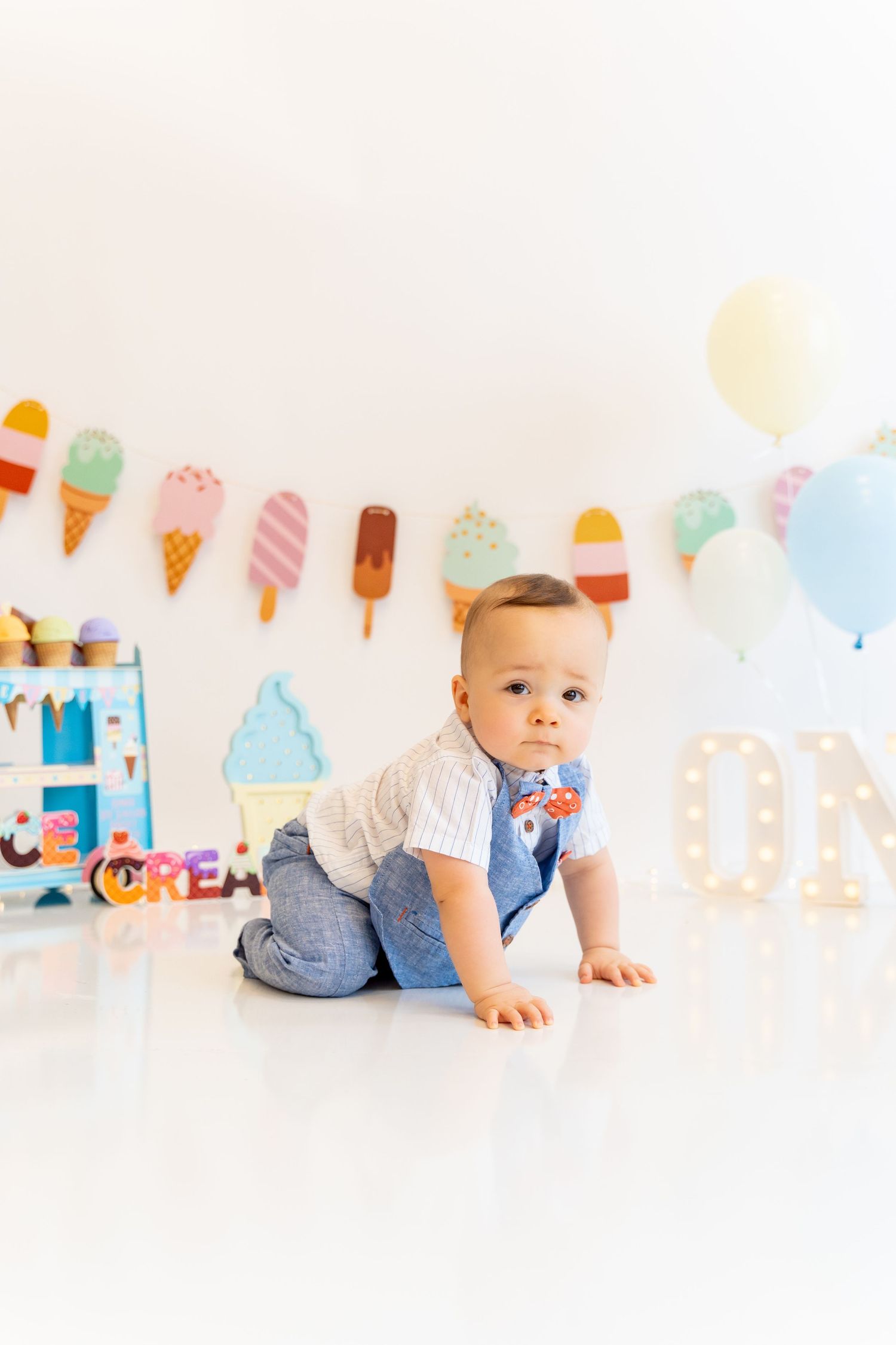 Baby in blue outfit crawling on white floor with colorful ice cream garland and party decorations.