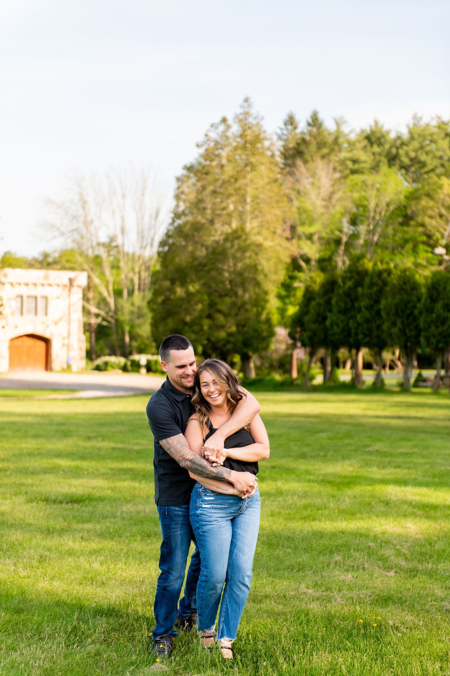Playful couple embracing and laughing on green lawn with stone building in background.