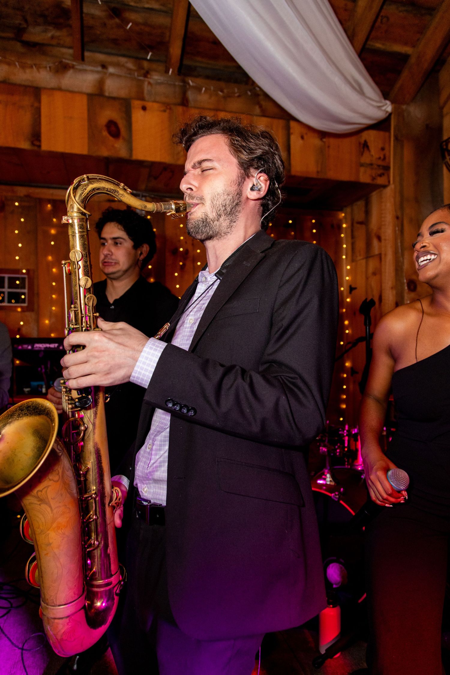 Musicians perform with saxophone at rustic venue with string lights and wooden beams.