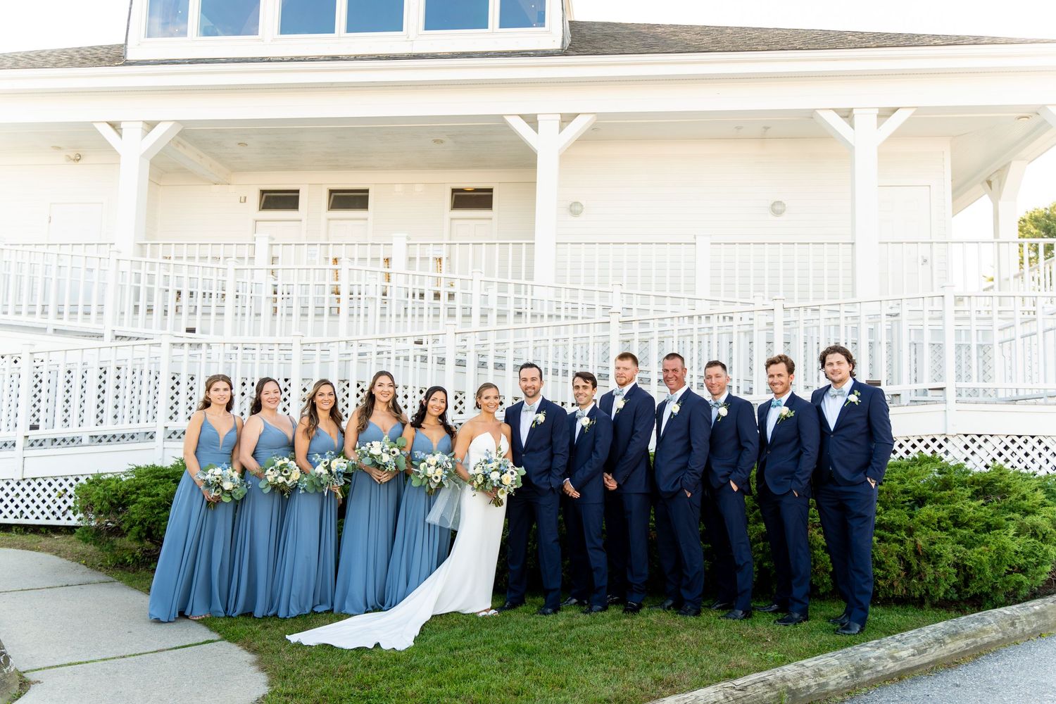 Wedding party in blue dresses and navy suits poses on white porch stairs.