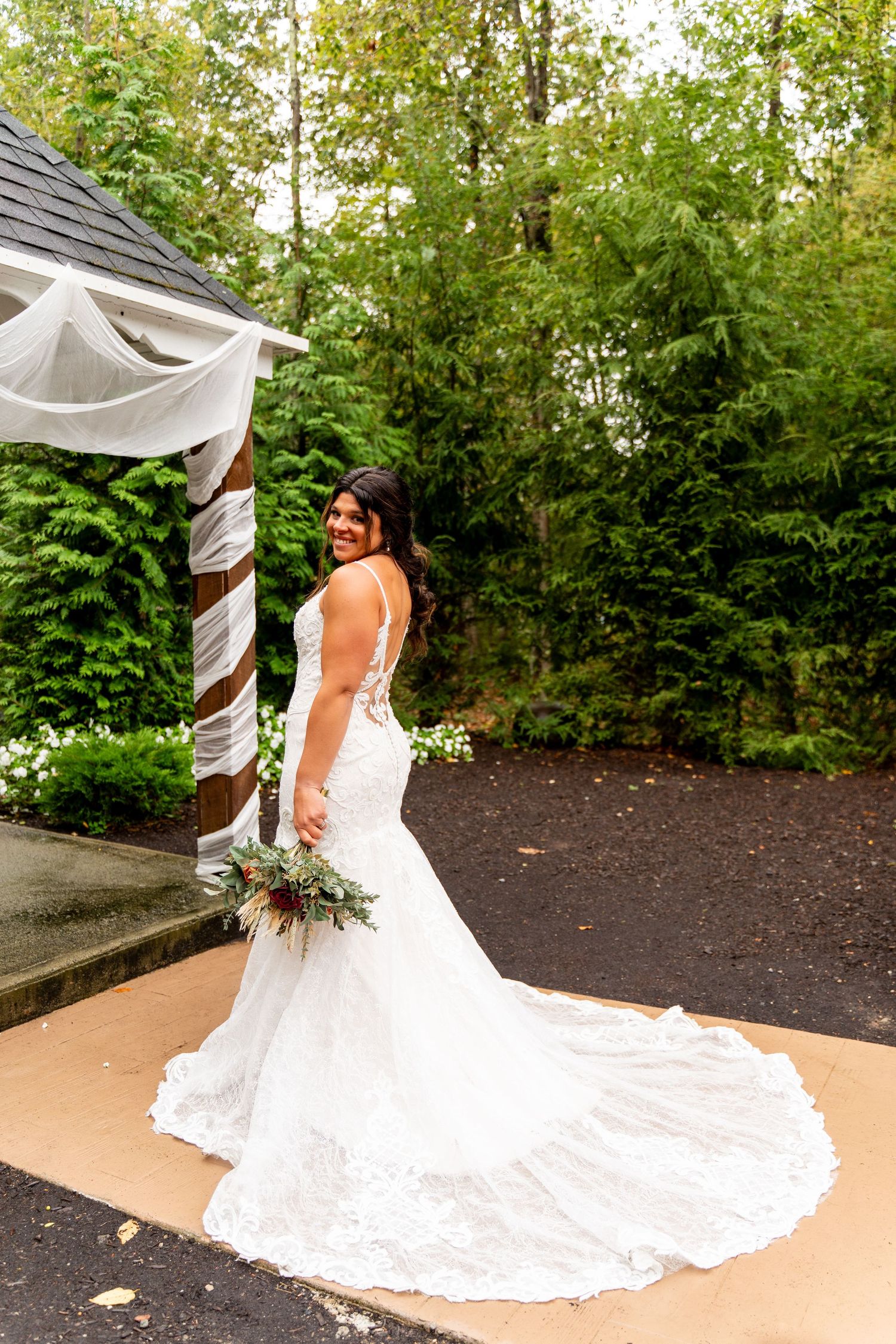 Bride in lace wedding dress with long train poses under outdoor gazebo.