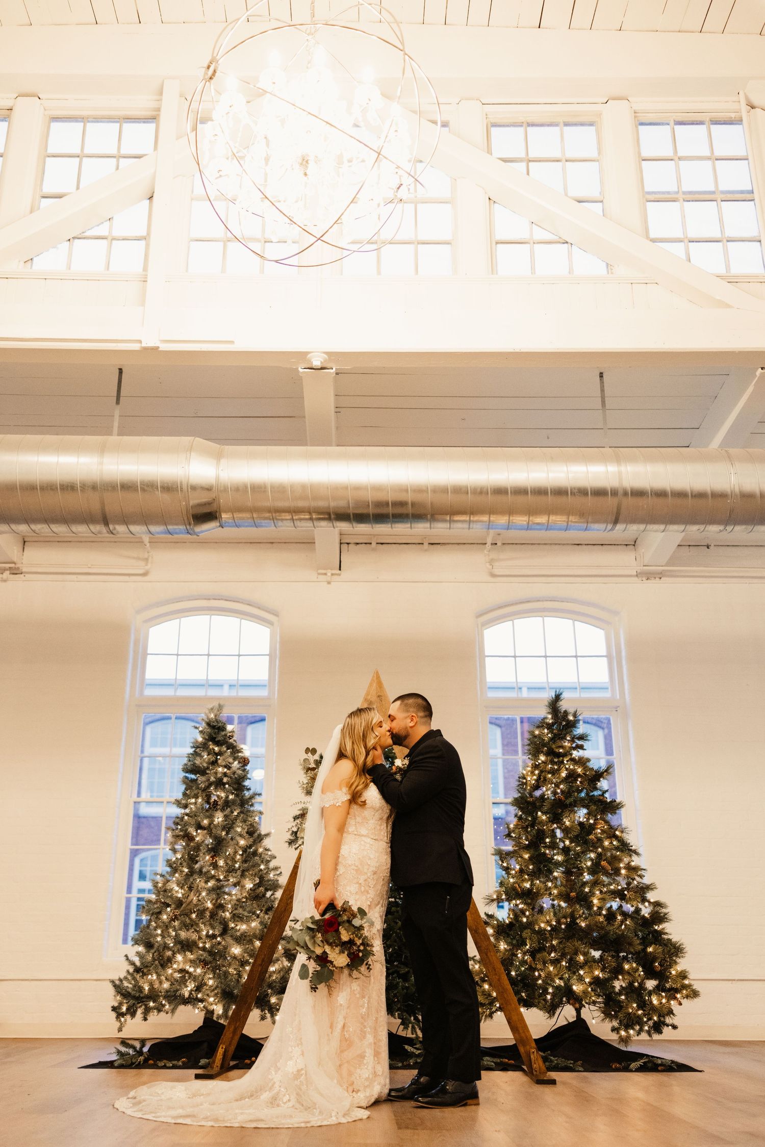 Wedding couple shares intimate moment between lit Christmas trees in bright industrial venue.