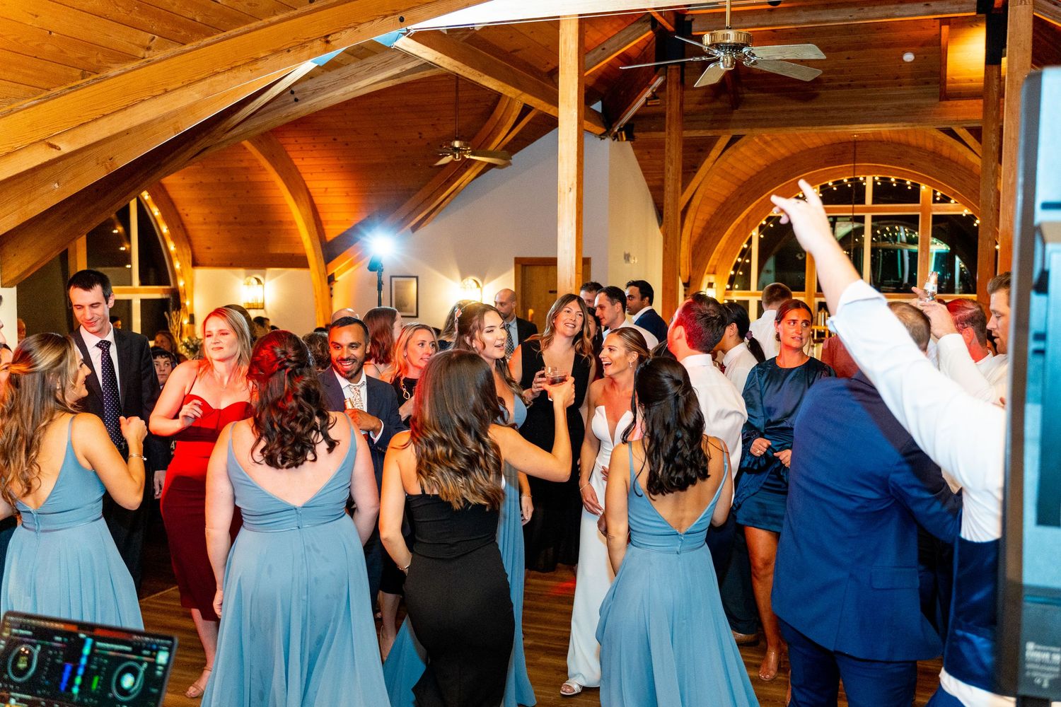 Lively wedding reception dance floor scene with guests celebrating under wooden beams.