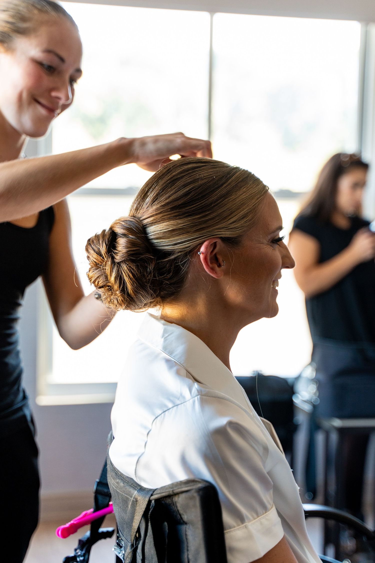 Hairstylist pins up elegant updo on client wearing white robe during wedding preparation.