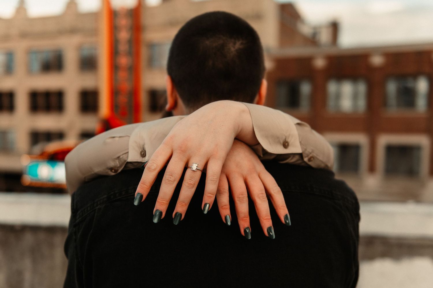 Close-up of engagement ring shown as couple embraces with city backdrop.