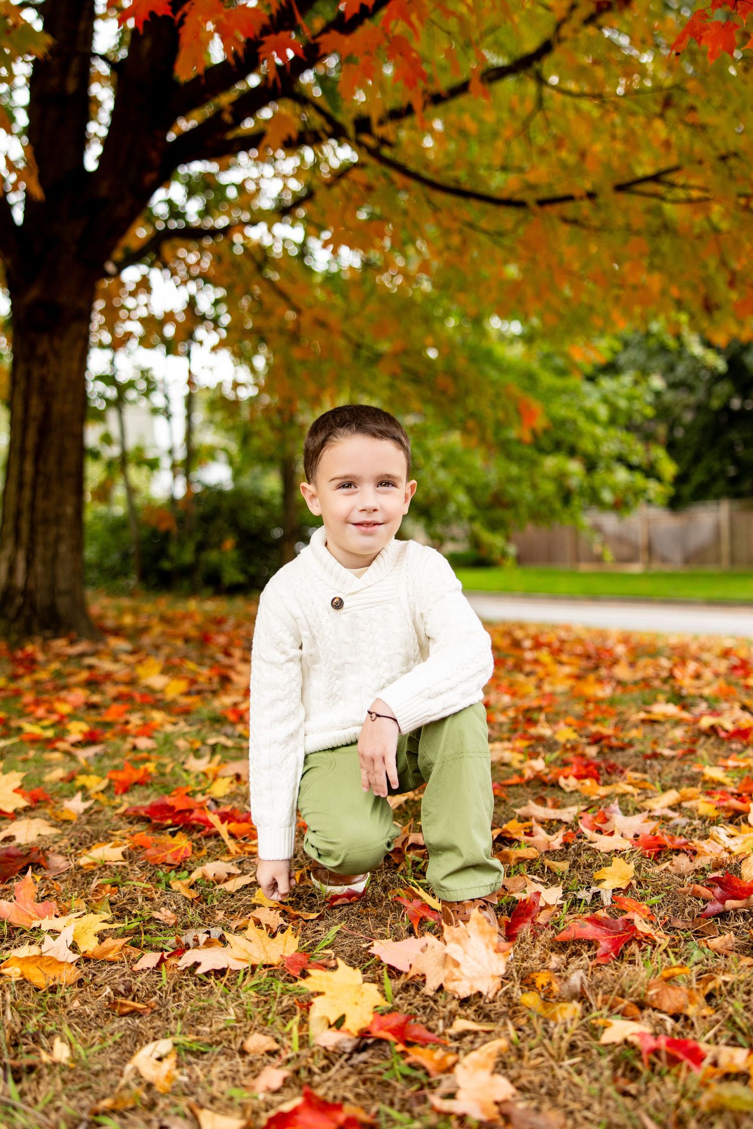 Young child crouching among colorful autumn leaves in a white sweater and khaki pants.