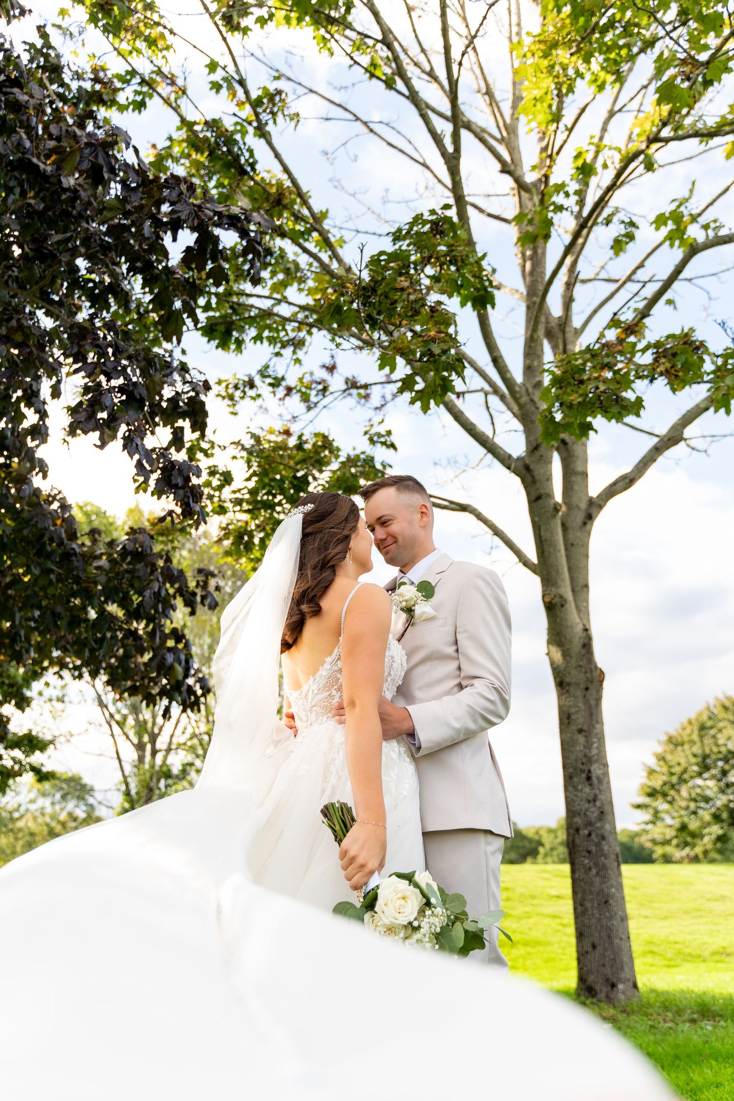 Wedding couple in formal attire photographed under tall trees on a grassy hill.