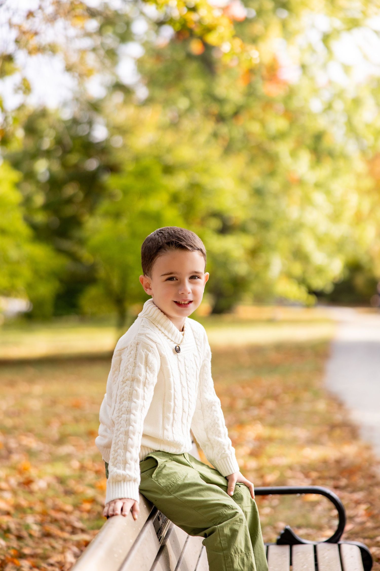 Young boy in cream sweater sitting on bench surrounded by autumn foliage.