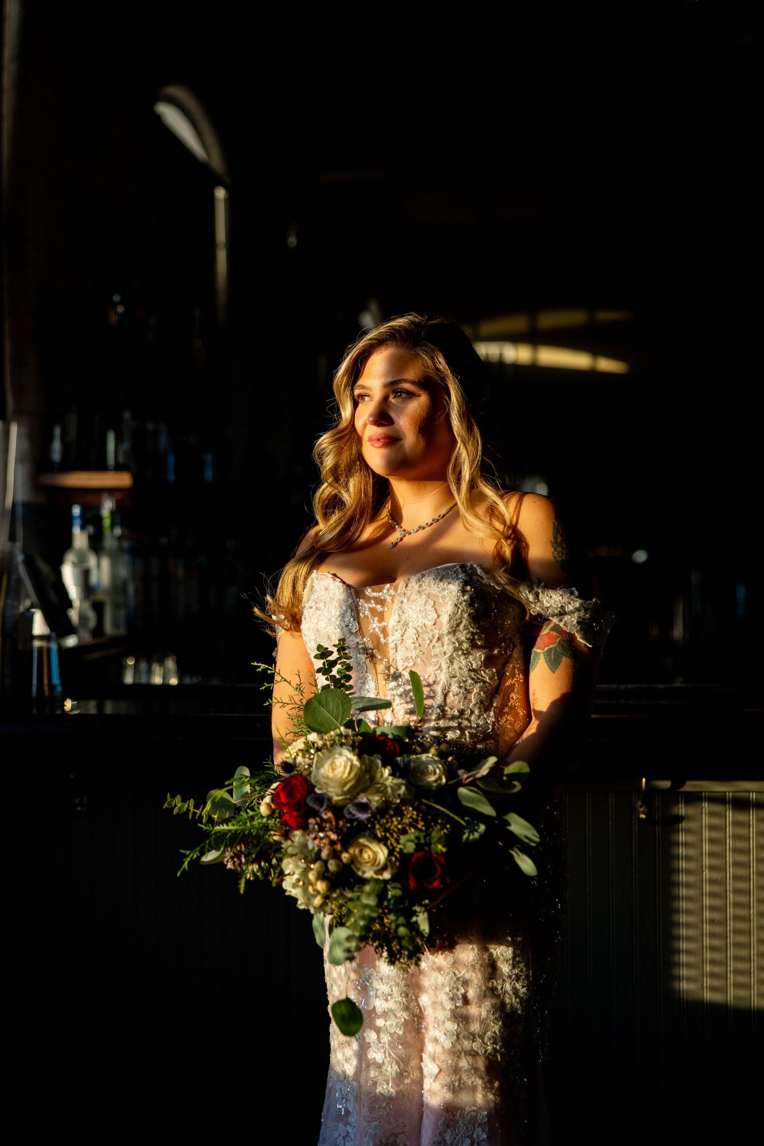 Bride in lace dress holds colorful bouquet while standing in dramatic lighting.