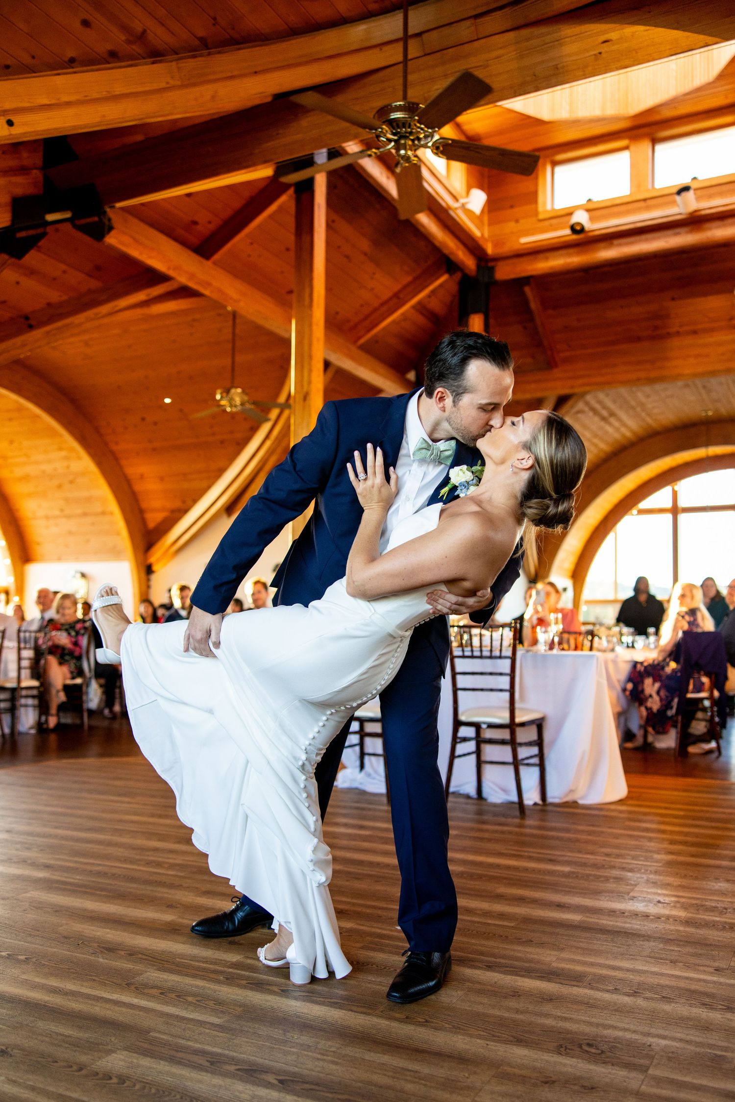 Newlyweds perform romantic dip dance move on wooden floor under vaulted ceiling.