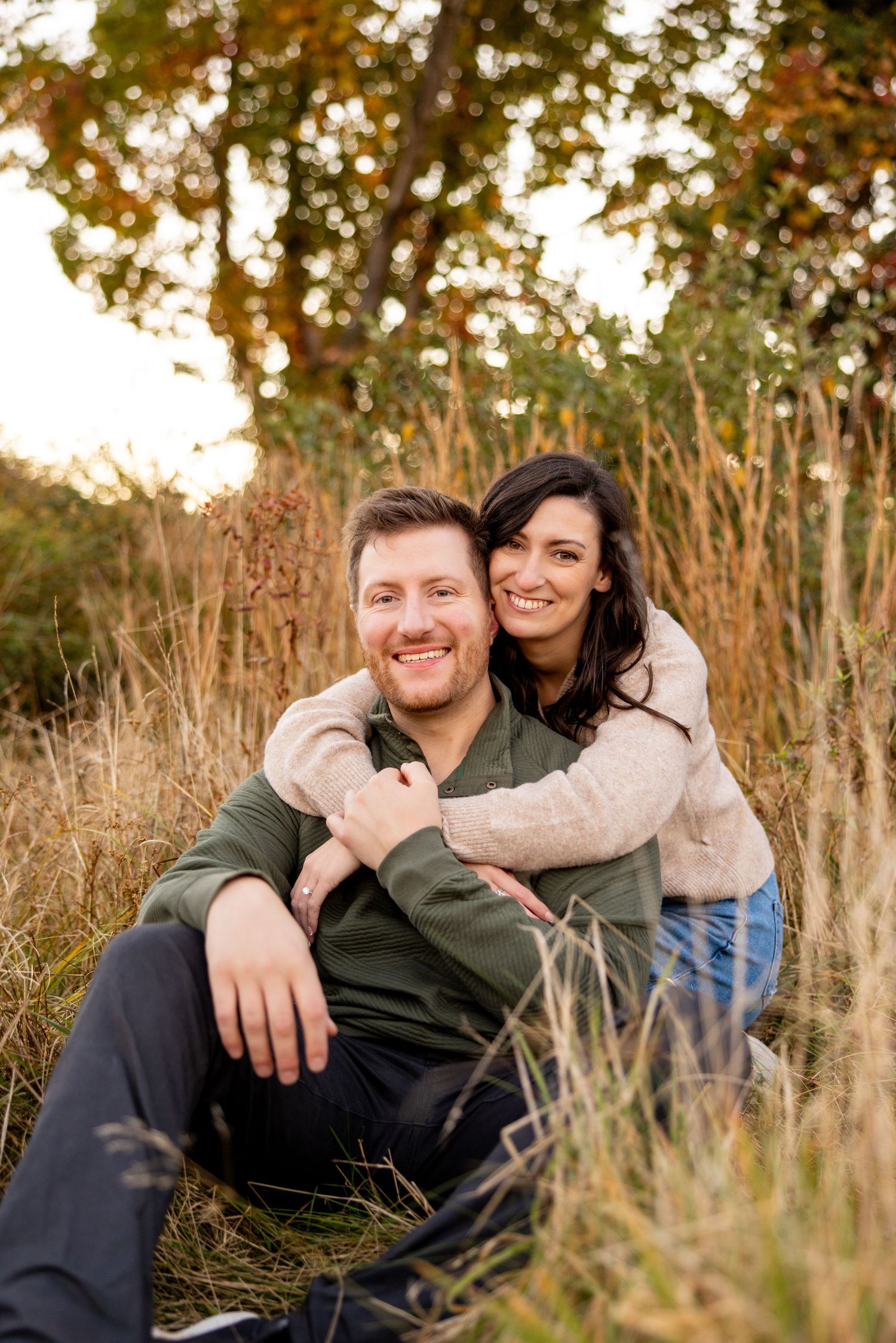 A couple sitting in tall grass during sunset, sharing a warm embrace.