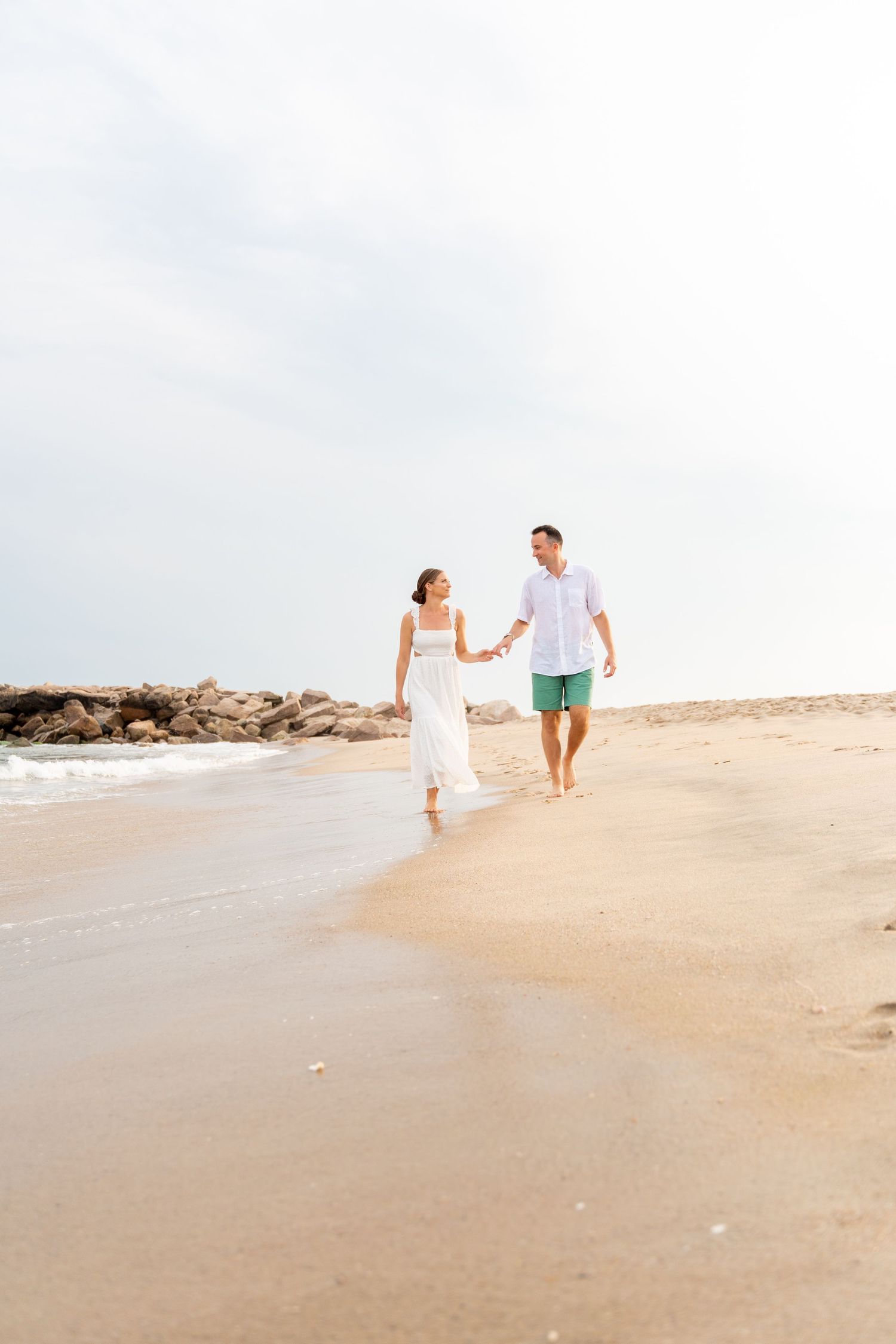 A couple walks hand in hand along a sandy beach with water and rocky jetty in background.