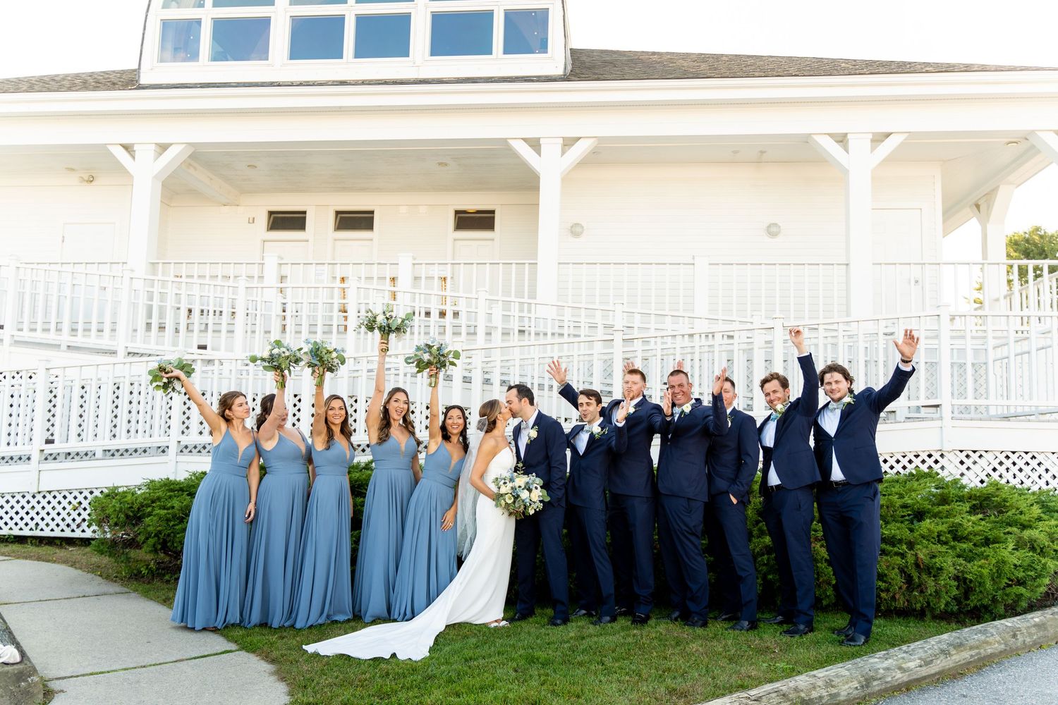 Wedding party in blue bridesmaid dresses and navy suits poses cheerfully in front of white building with porch railings.