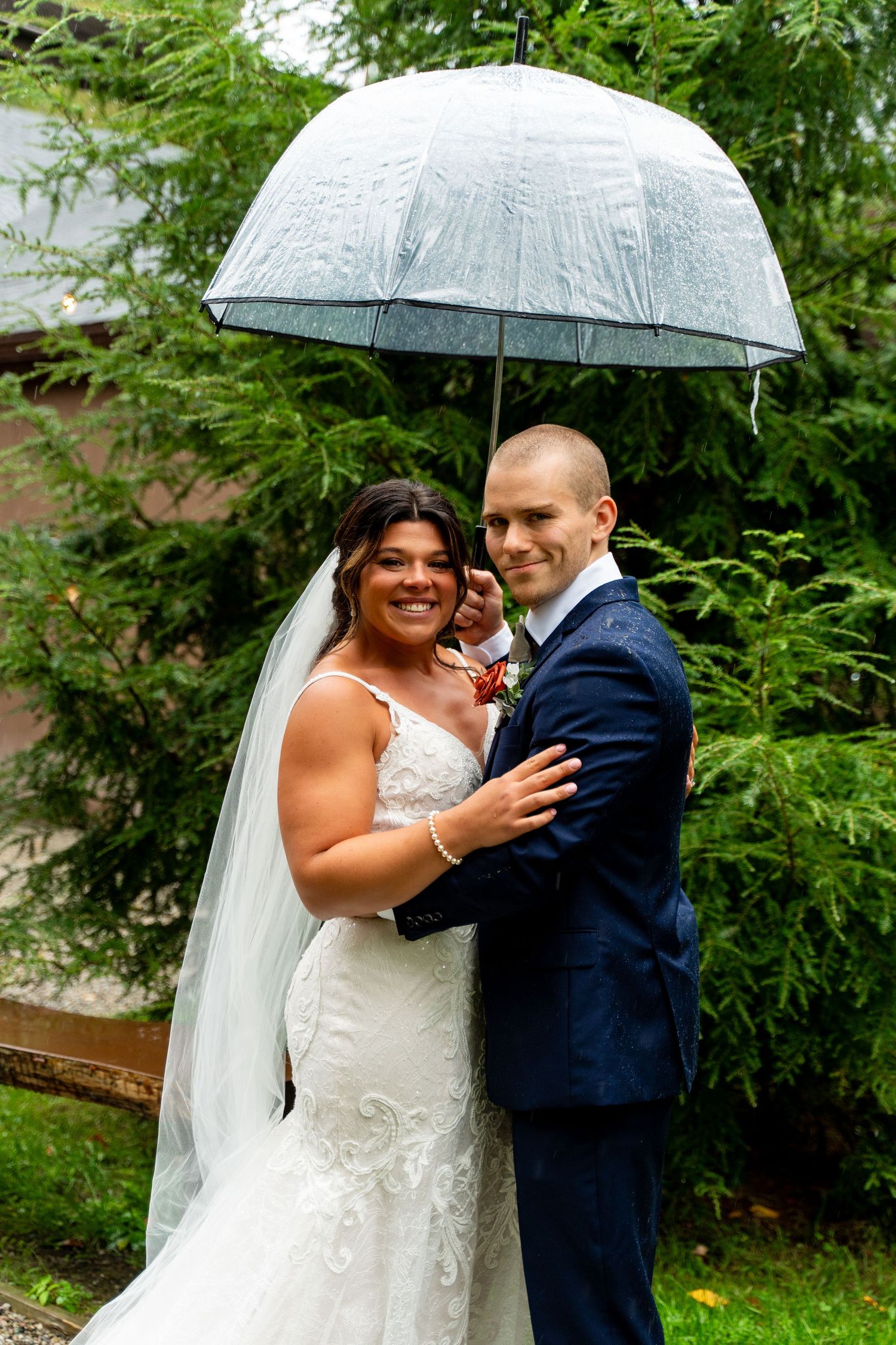 A happy couple embraces under a clear umbrella in a lush garden setting on their wedding day.