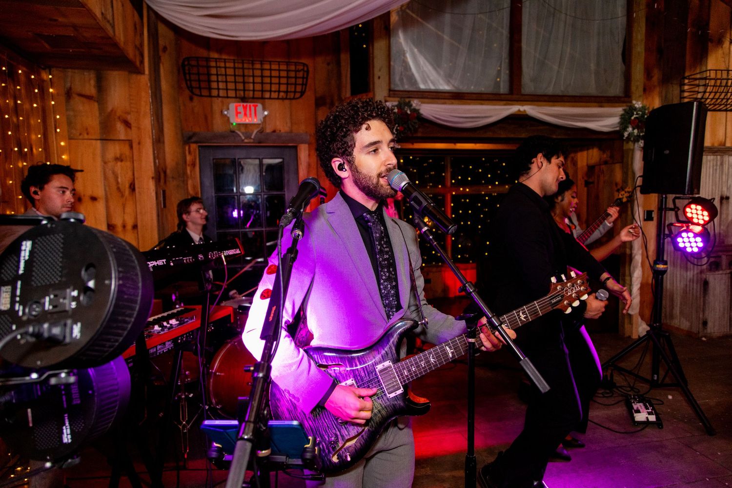 Band member plays guitar during performance in rustic barn venue with purple lighting.