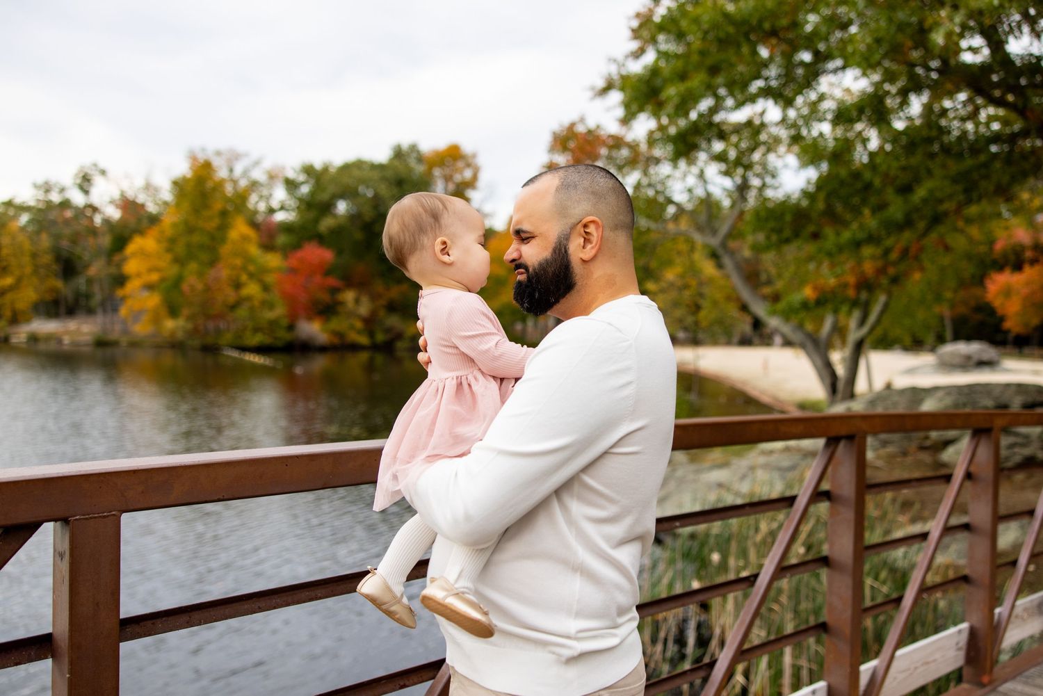 Parent holds baby while standing on wooden bridge overlooking autumn lake.