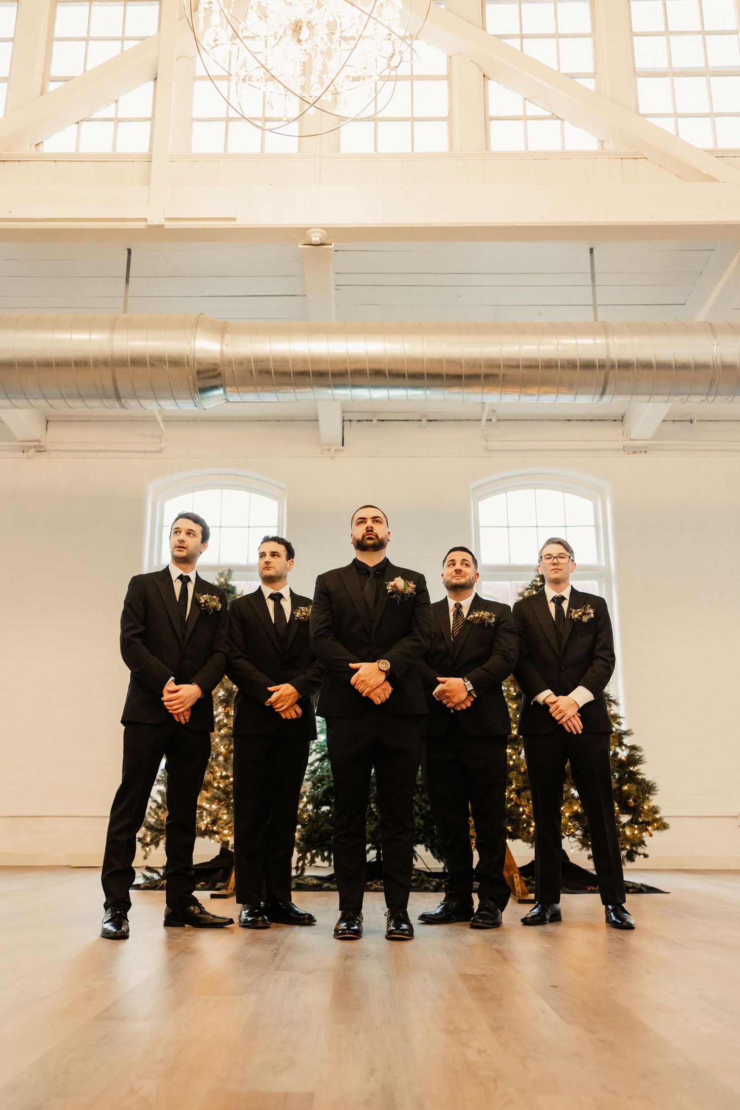 Groomsmen in black suits pose formally in bright industrial space with high windows.
