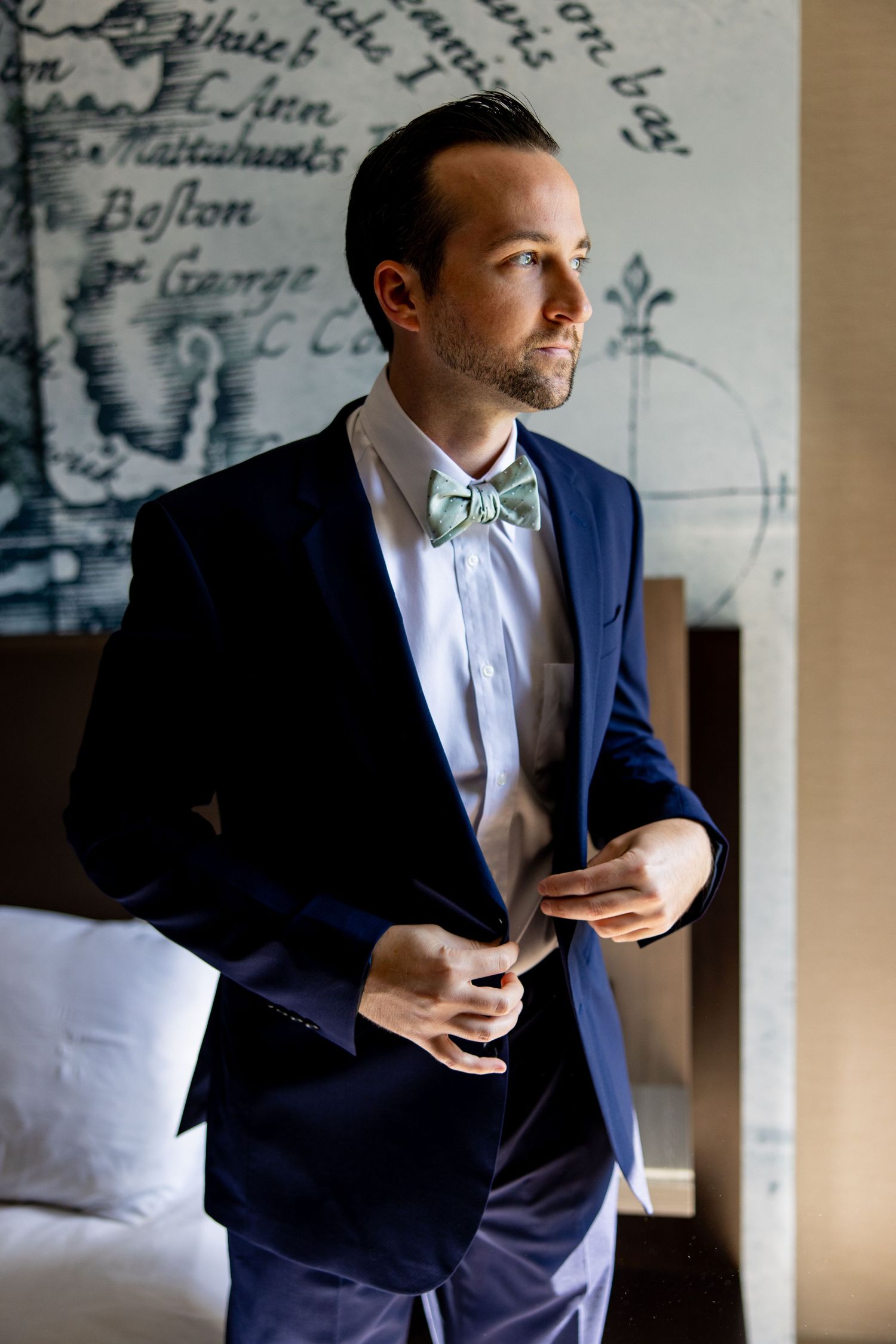 Groom in navy suit and bow tie adjusts jacket while standing against patterned wall.